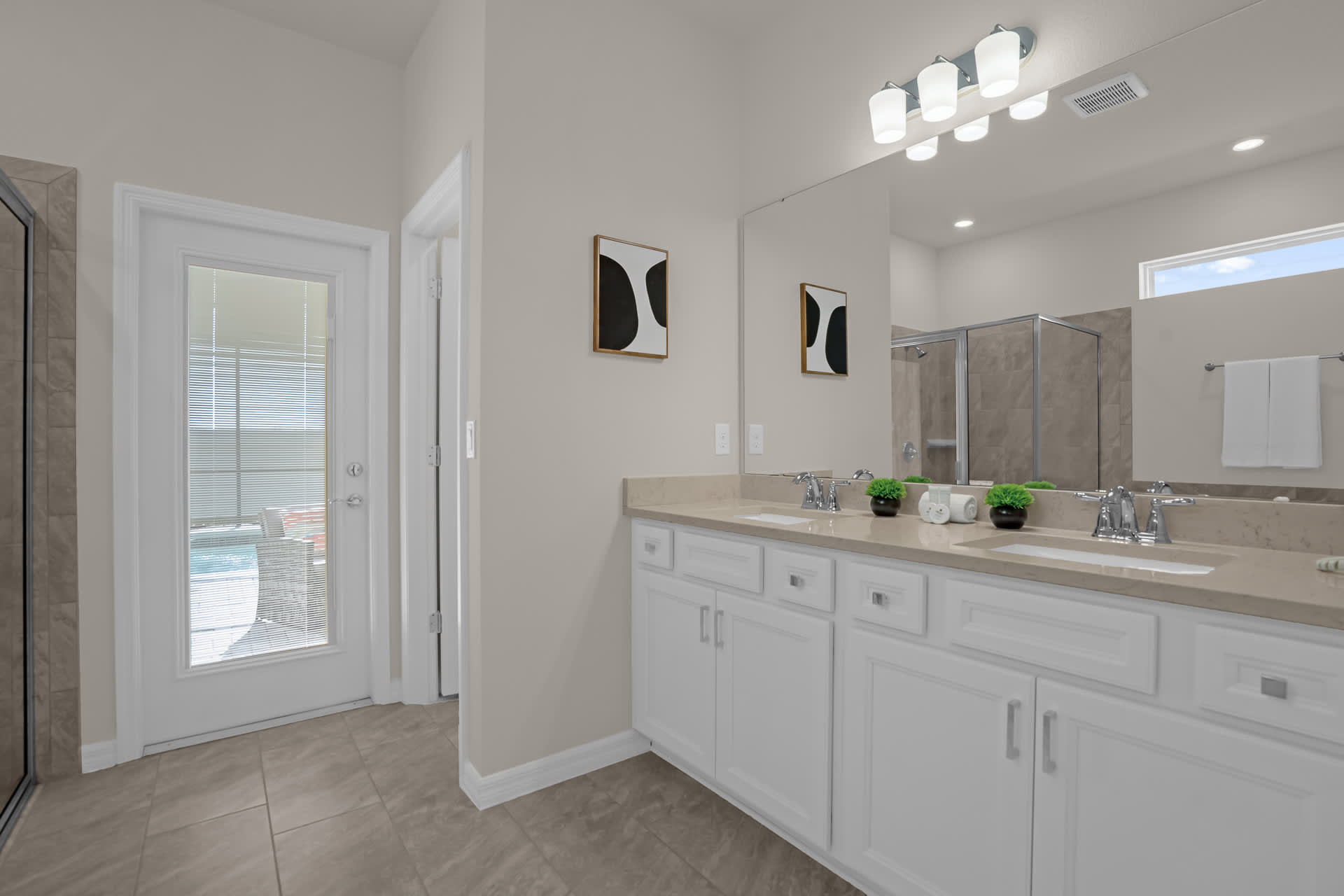 Bright bathroom with a double vanity featuring white cabinets, light-colored countertops, and a large mirror, with a door leading to an outdoor area.