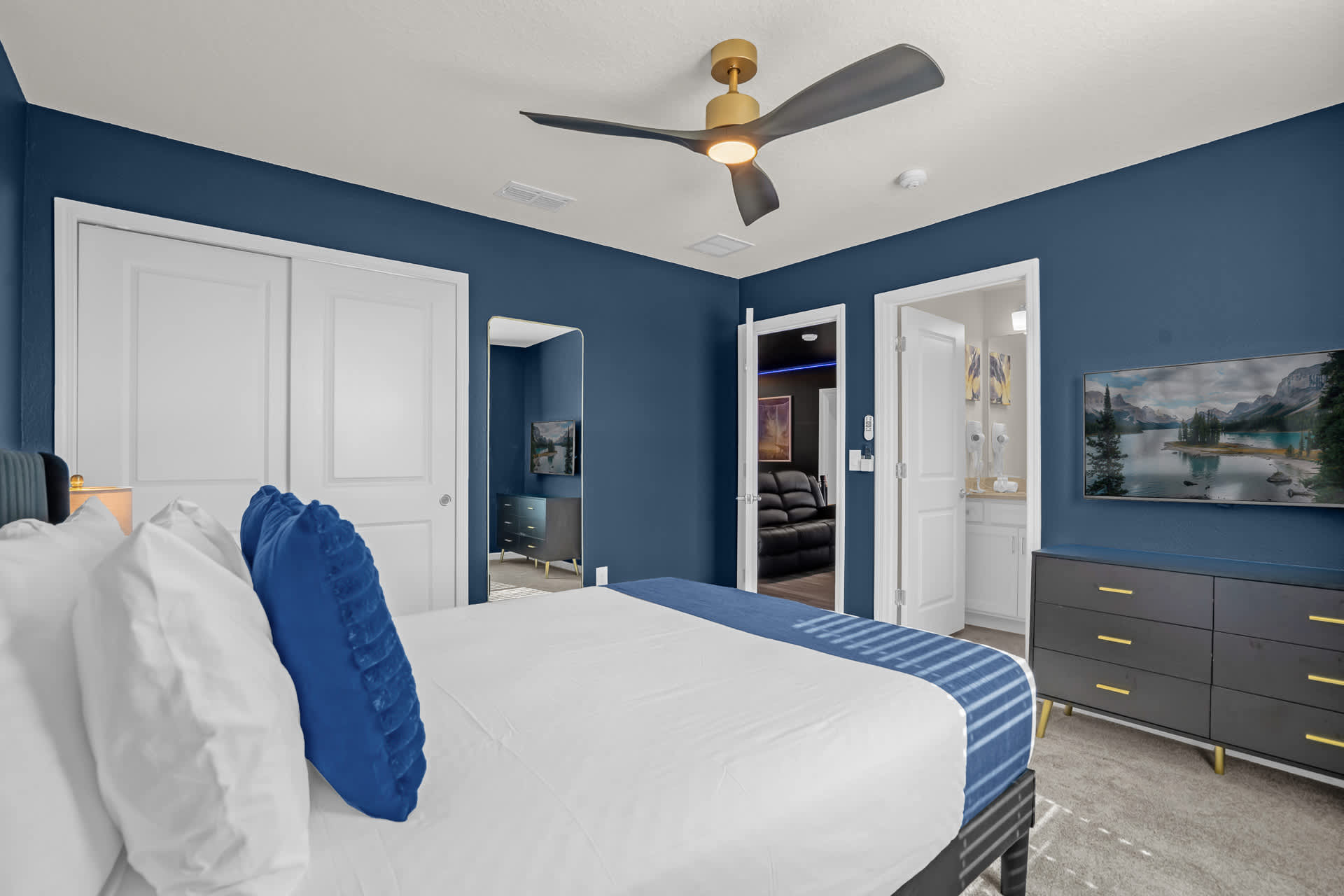 Another angle of the blue-themed bedroom, showcasing a dresser with a TV, a full-length mirror, and an open doorway leading to another room, with a modern ceiling fan overhead.