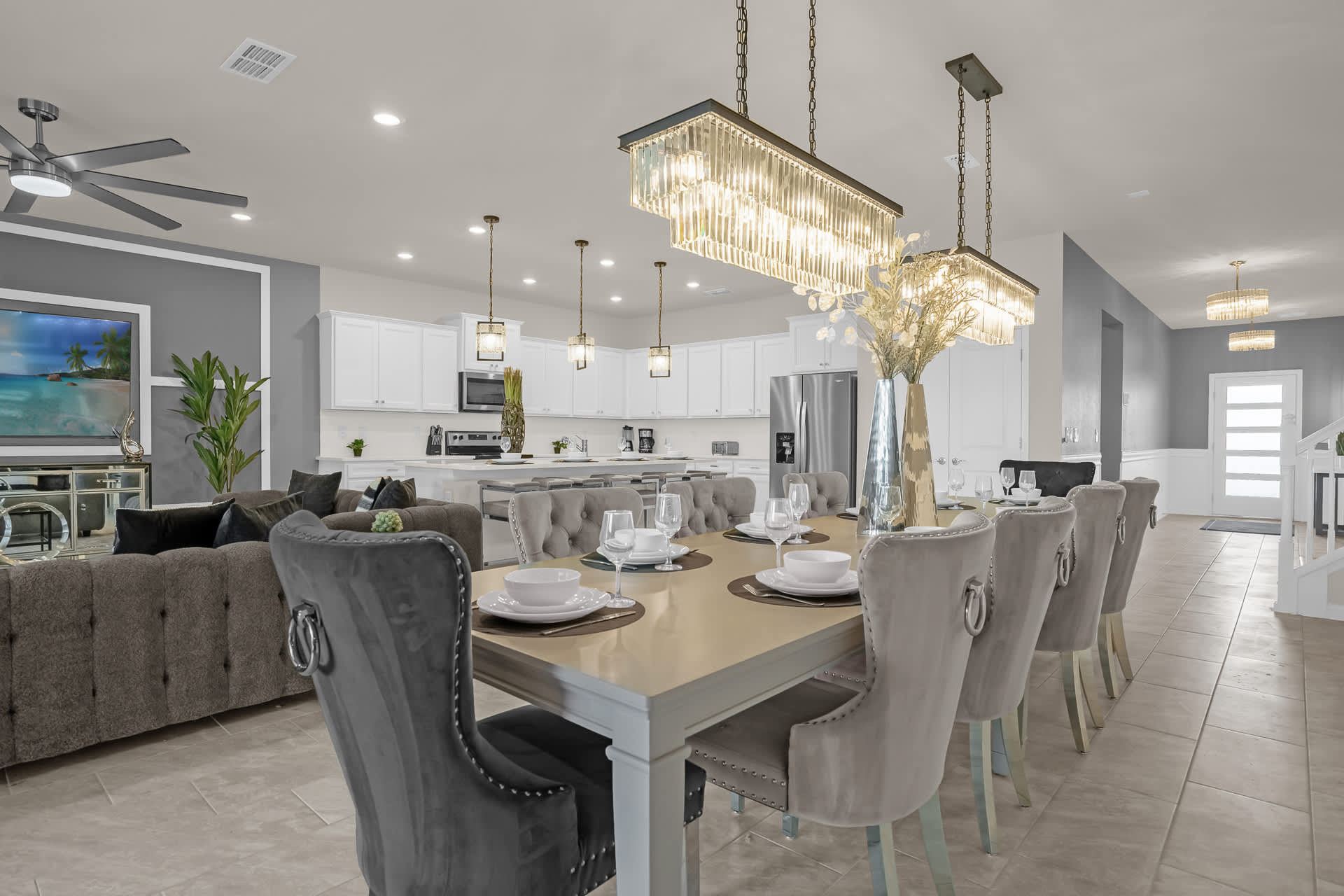 An elegant open-concept dining area with a long wooden table and upholstered gray chairs, illuminated by two rectangular crystal chandeliers, adjacent to a modern kitchen and living room.