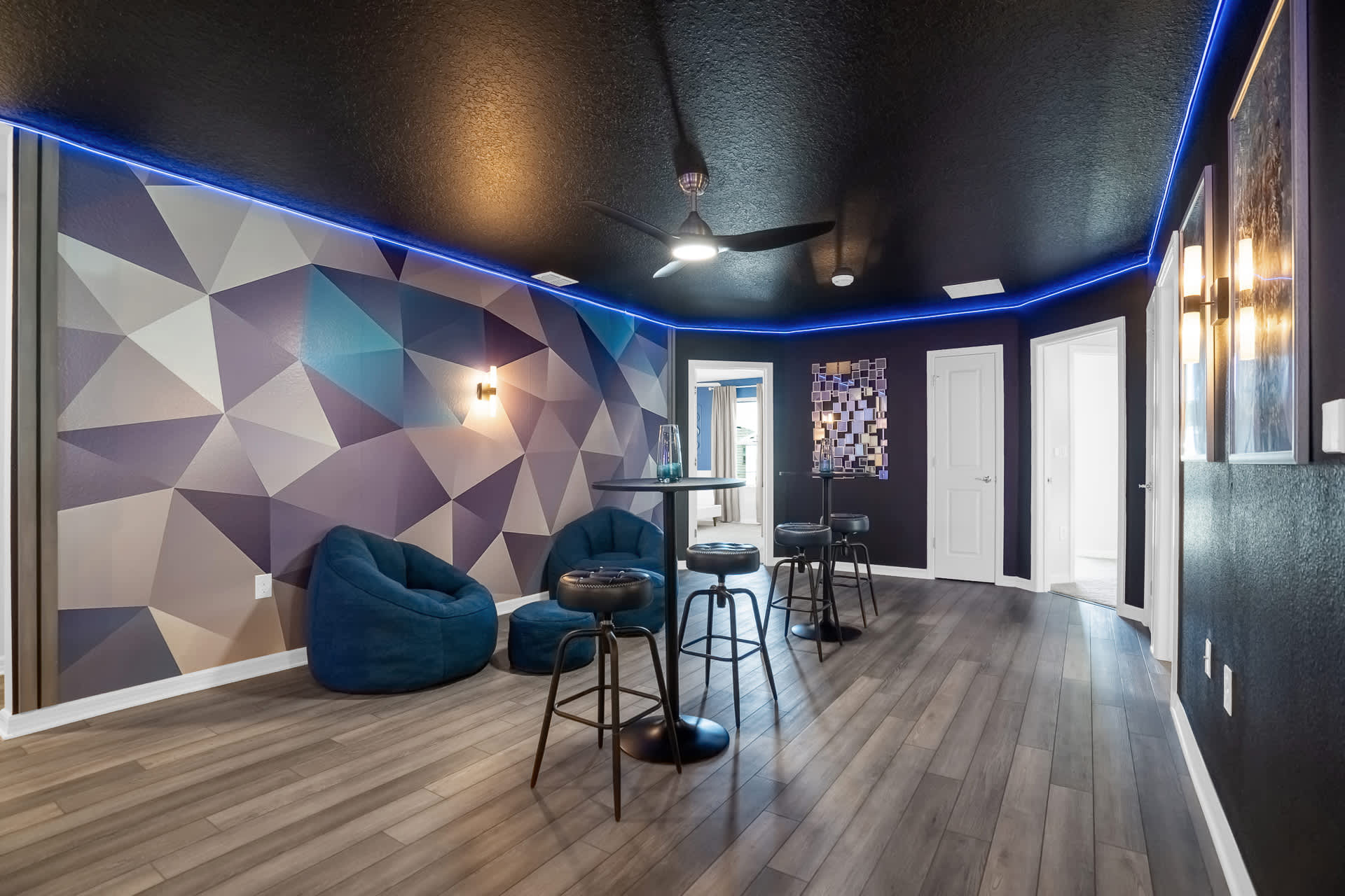 A modern lounge area with a striking geometric accent wall, two blue bean bag chairs, and two high-top tables with black stools, featuring a ceiling fan and blue LED strip lighting.