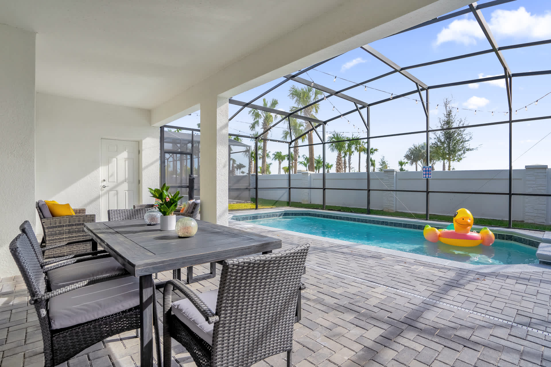 Covered outdoor patio area with a dark gray dining table and chairs, lounge seating, and a BBQ grill, adjacent to a screened private pool with an inflatable duck float.