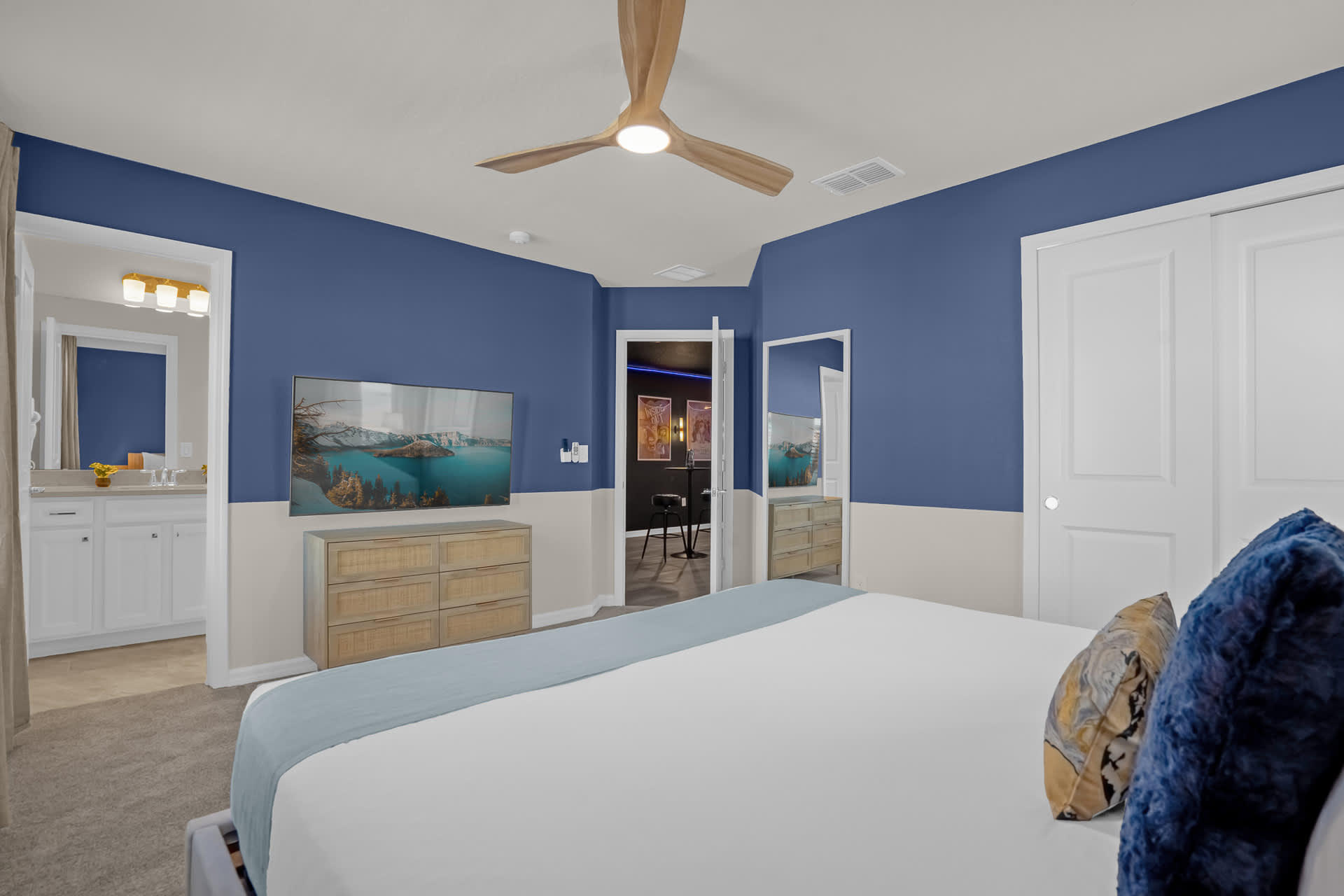 View of the bedroom showing a large flat-screen TV on a wooden dresser, a full-length mirror, and a doorway leading to an adjacent bathroom.