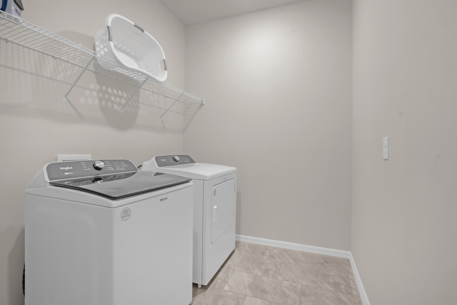 A functional laundry room equipped with a white top-load washing machine and a matching dryer, with a wire shelf installed above for storage.