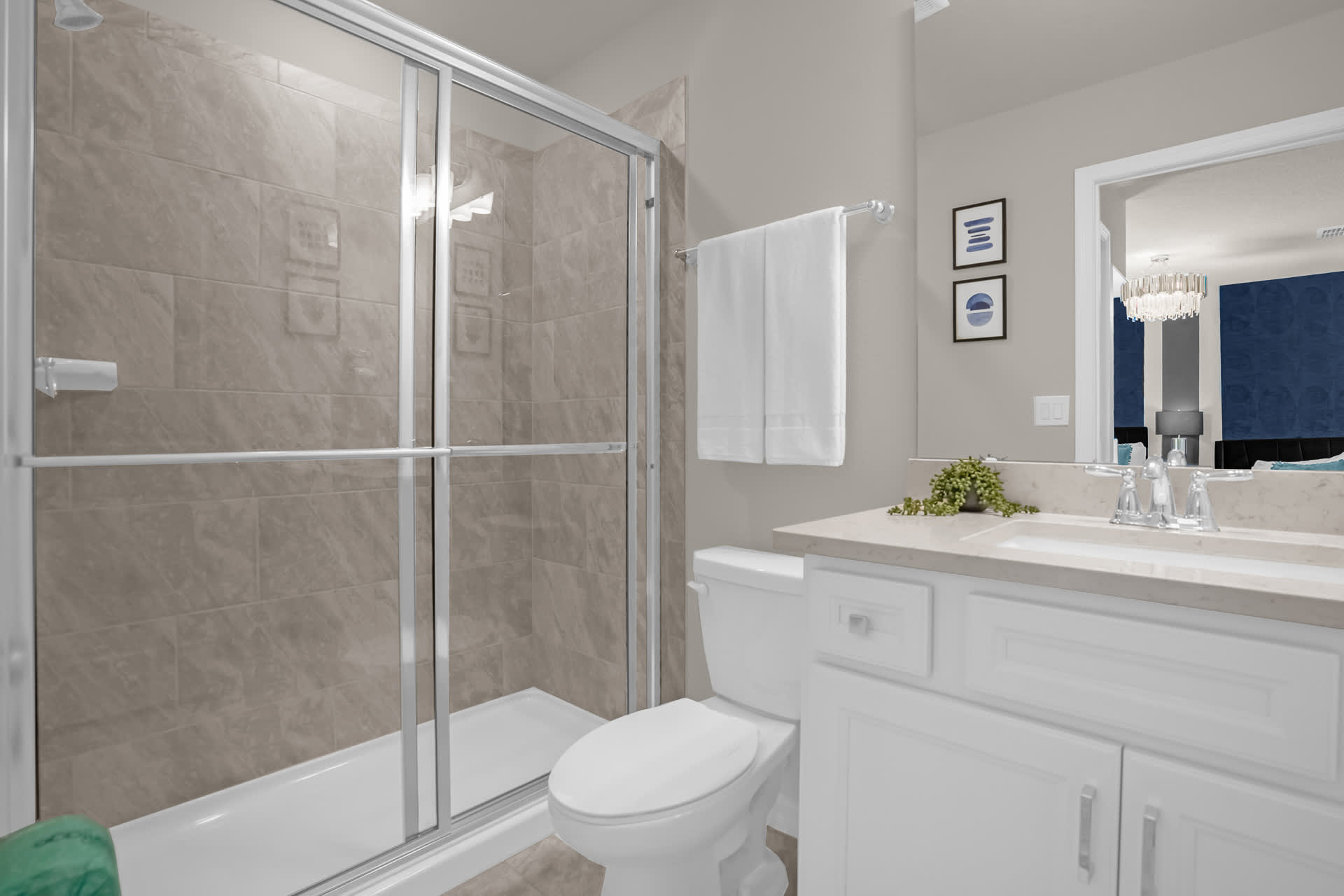 A well-lit bathroom featuring a glass-enclosed walk-in shower with light-colored tiles, a white vanity with a single sink, and a toilet, with minimalist wall art above the toilet.