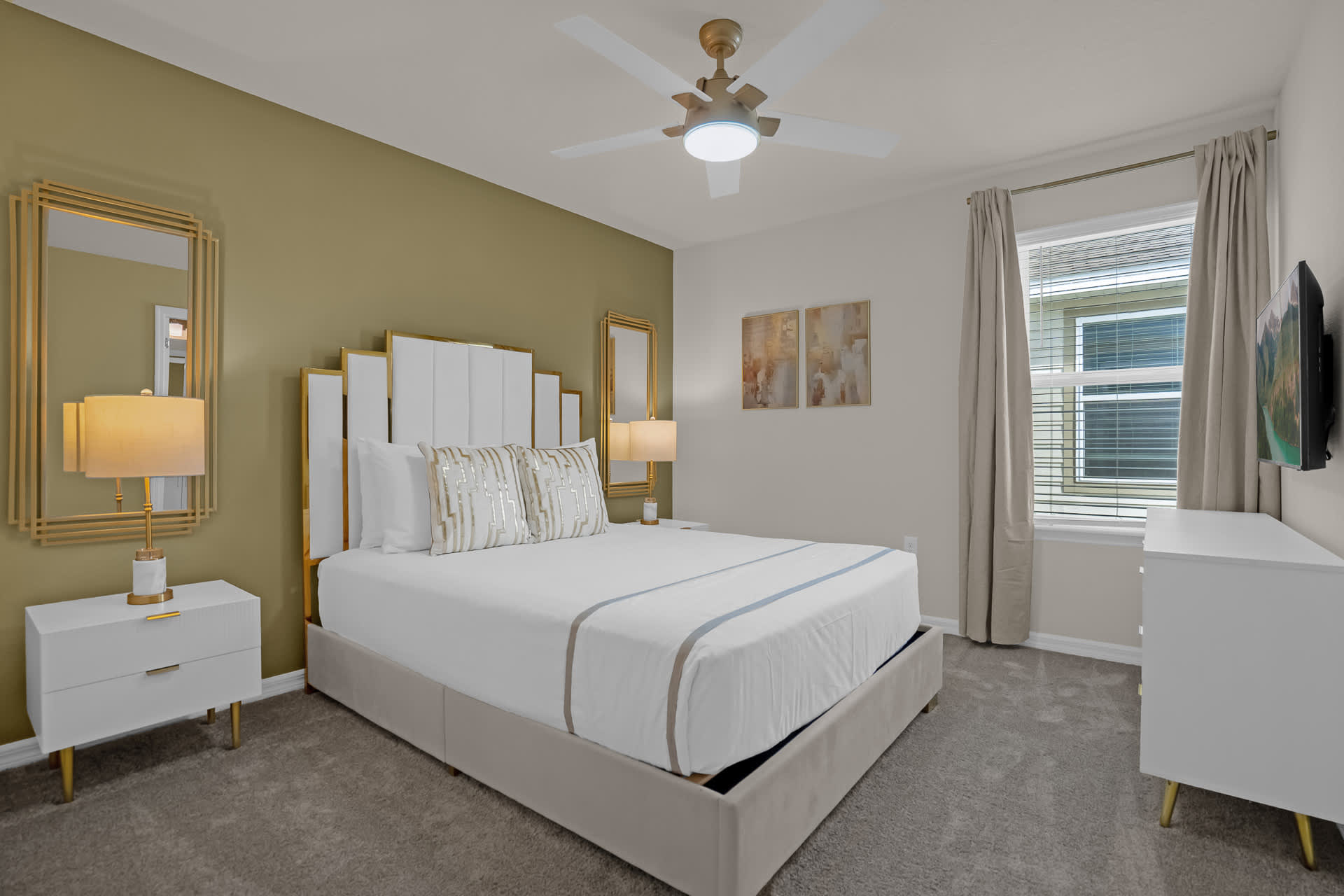 A spacious bedroom with a king-sized bed featuring a tall white and gold headboard, two white nightstands with lamps, and a ceiling fan, set against a two-tone wall with a green accent.
