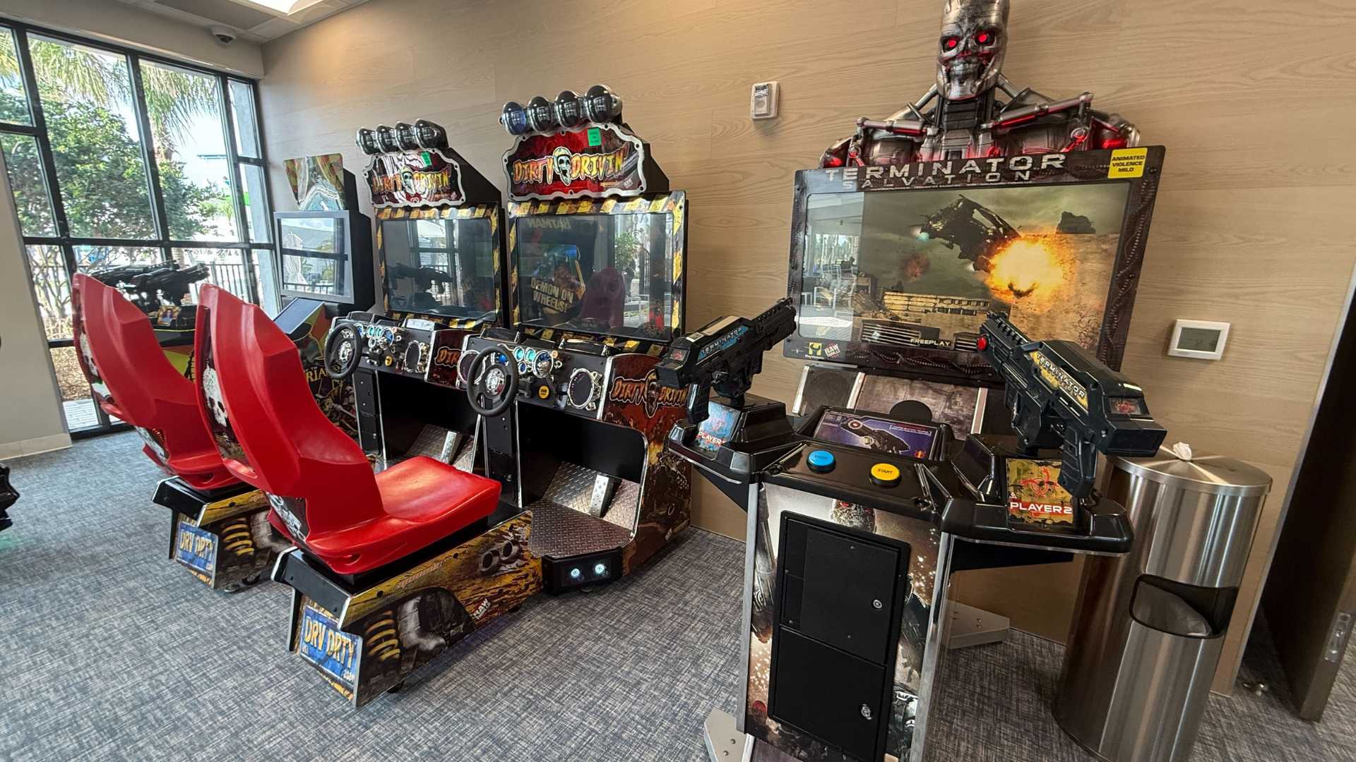 A section of the arcade room showcases several driving simulation arcade games with red seats and a "Terminator Salvation" shooting game, set against light wood paneling and carpeted flooring.