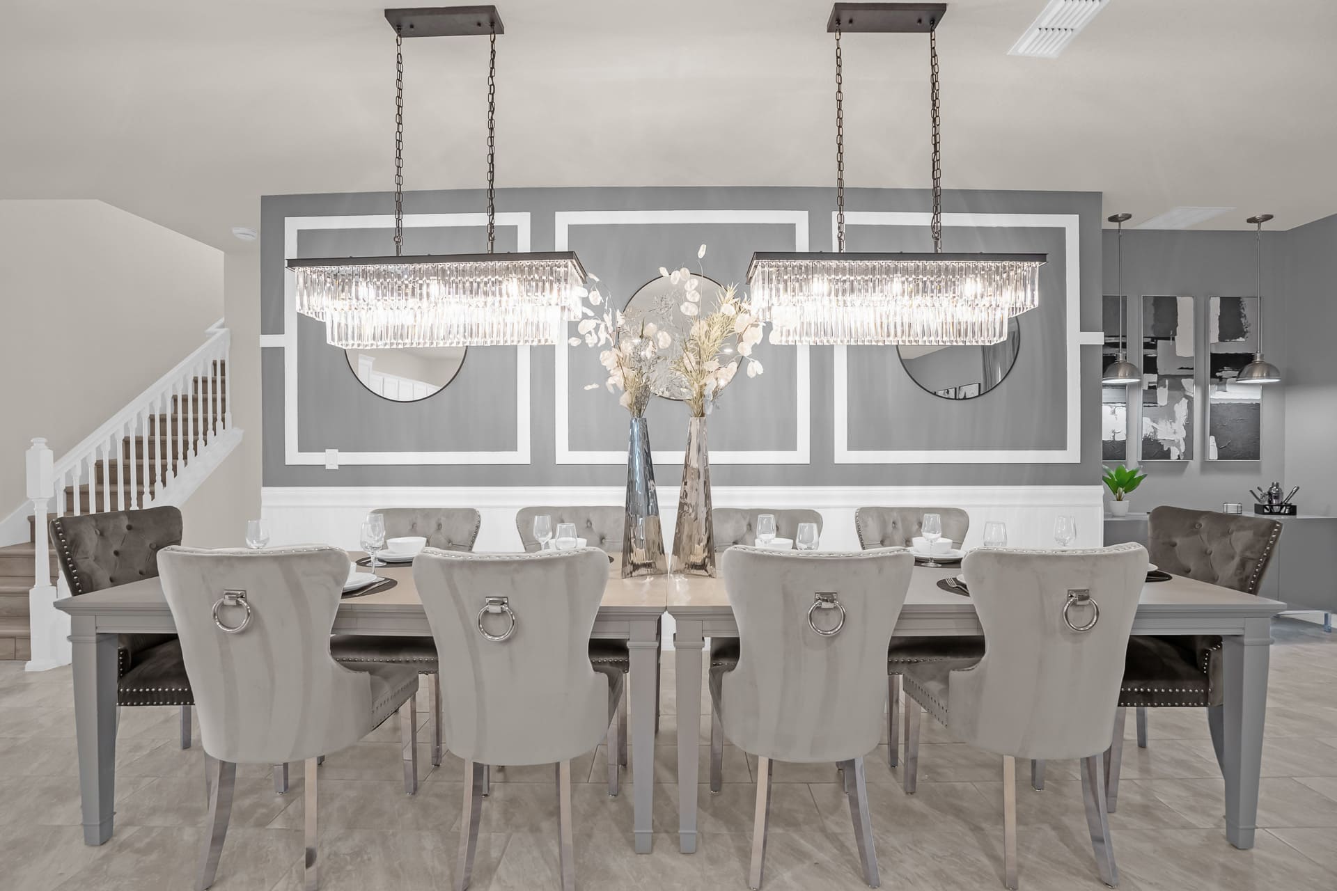 A sophisticated dining area featuring a long light-colored dining table with gray upholstered chairs, two ornate crystal chandeliers, and a gray accent wall with decorative mirrors.