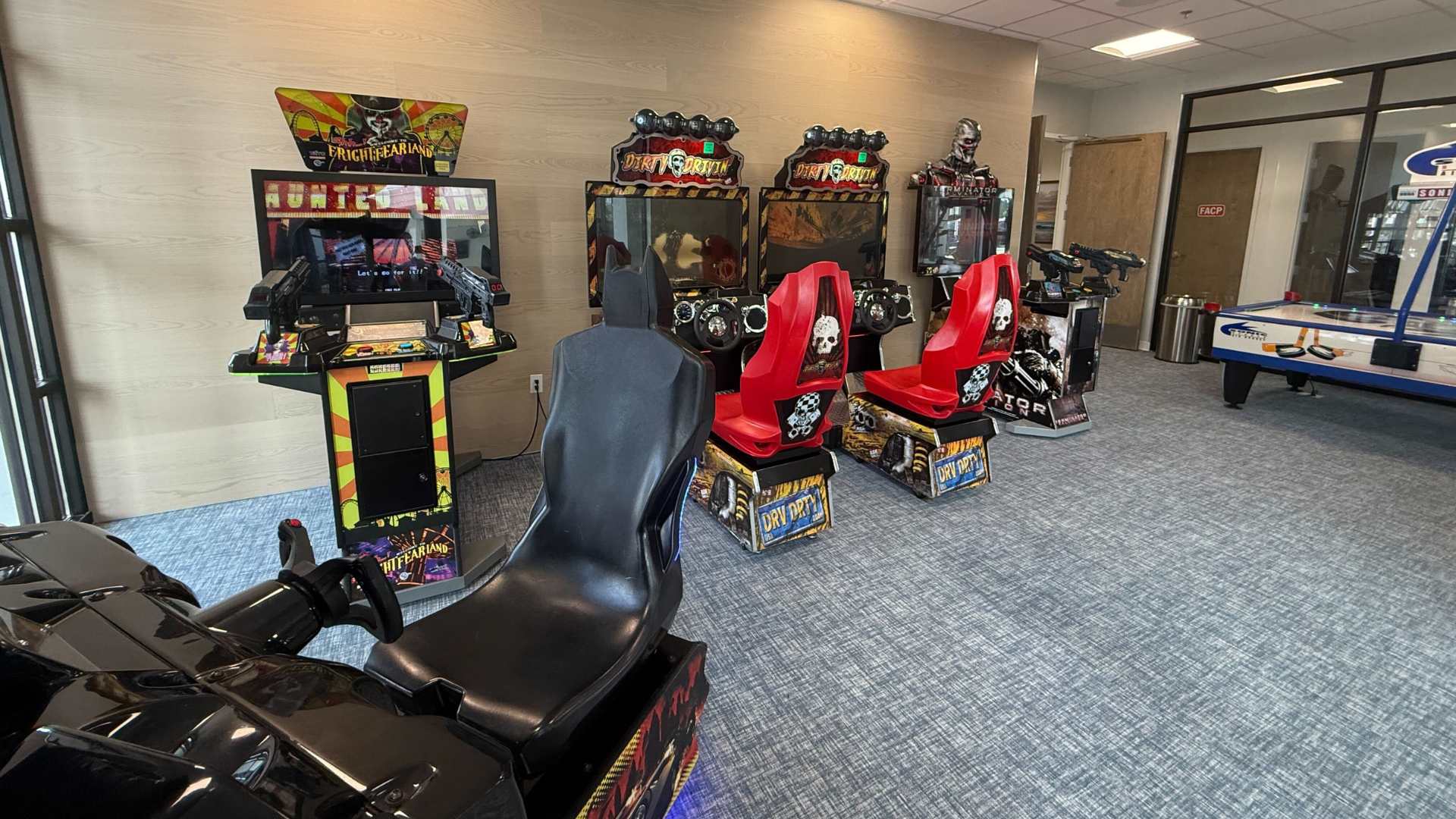 A wide shot of a modern game room showcases various arcade machines, including a Batman racing game, Dirty Driving, and Terminator, set on a gray patterned carpet. An air hockey table is visible in the background, complementing the light wood-paneled walls.