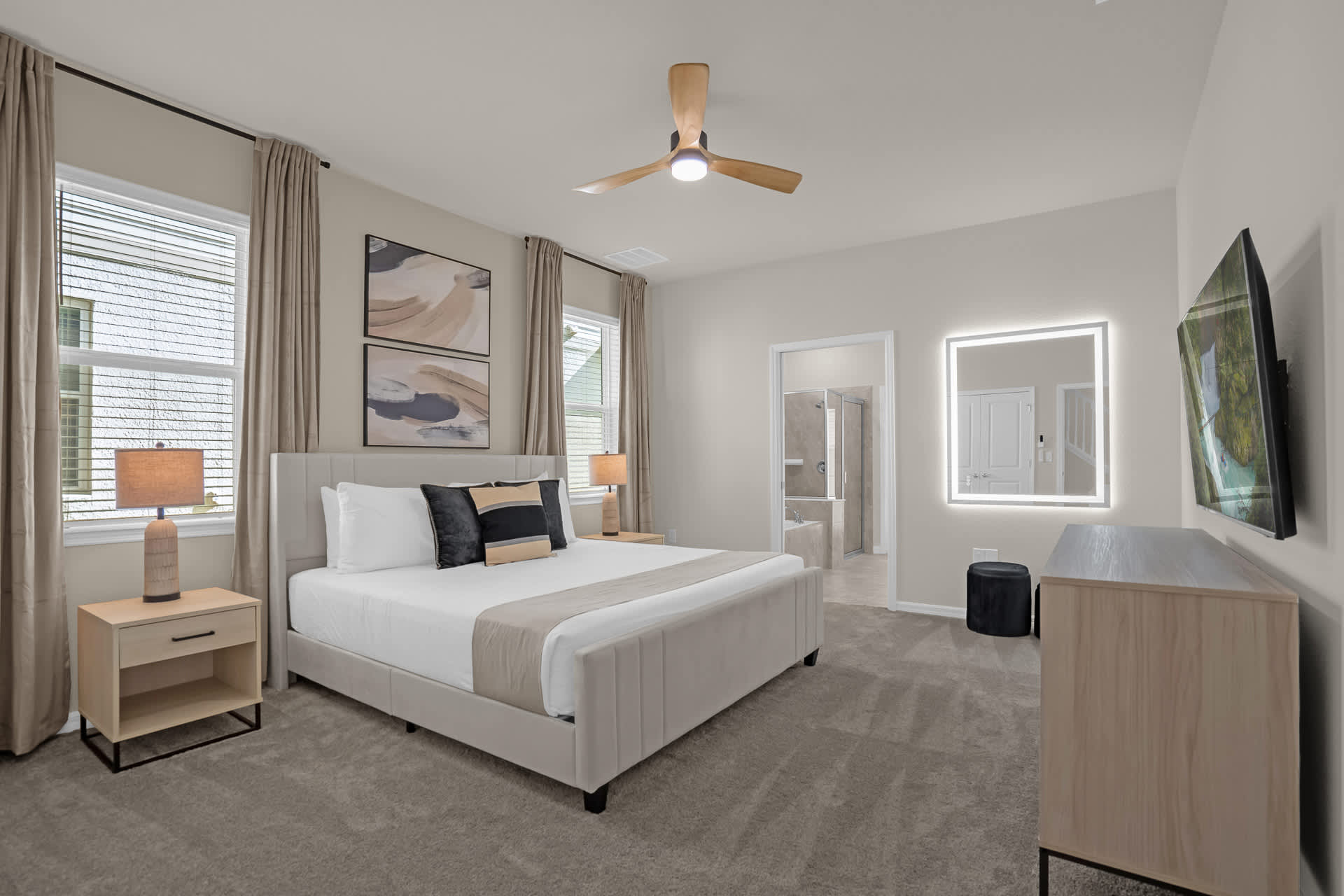 Spacious master bedroom with a light gray upholstered king bed, two wooden nightstands with lamps, a ceiling fan, and two large windows with light brown curtains.