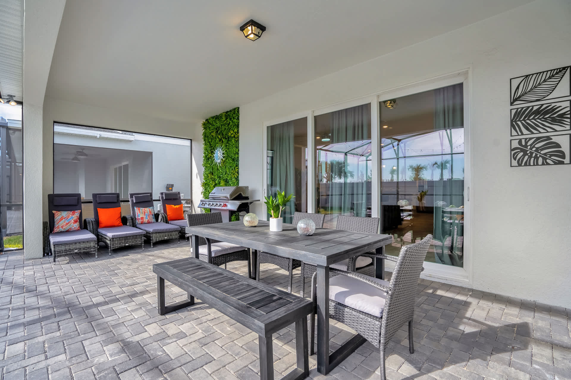 Spacious covered patio featuring a dark wood dining table with a bench and chairs, several lounge chairs, and a BBQ grill, all overlooking a screened private pool.