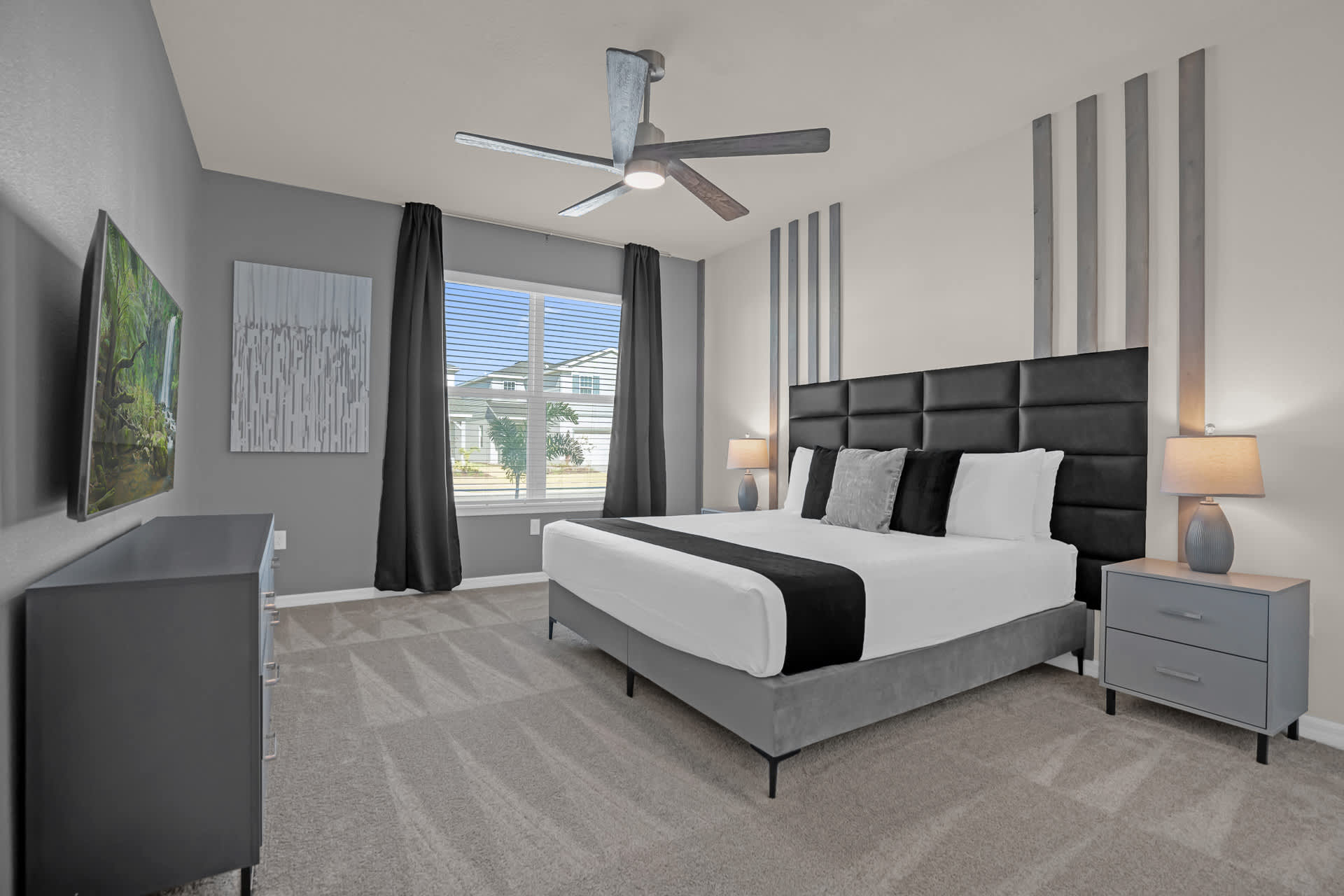 Spacious master bedroom featuring a king-sized bed with a black upholstered headboard, grey carpet, and a modern ceiling fan. Large windows with dark curtains provide natural light, complemented by bedside lamps and a dresser with a TV.