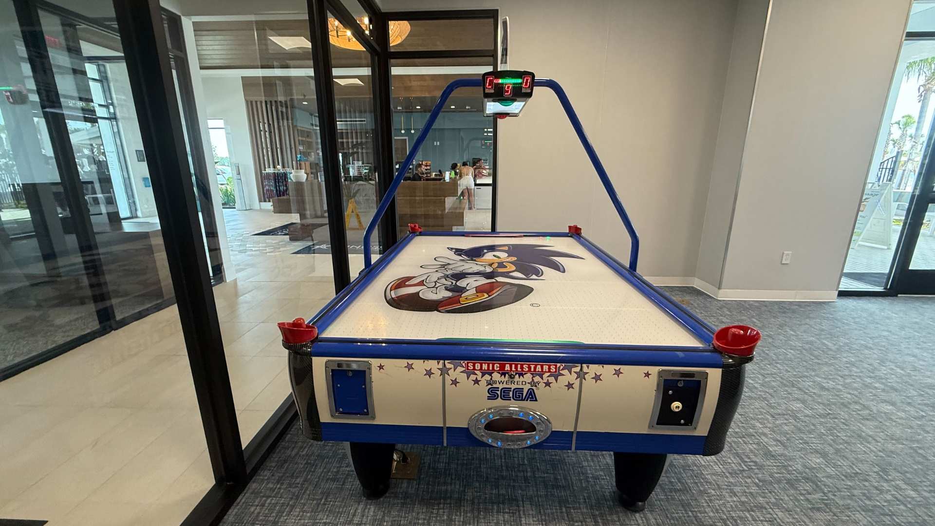 A close-up view of a "Sonic Allstars" air hockey table, featuring the character Sonic the Hedgehog, with a digital score display and a glimpse of other arcade games in the background.
