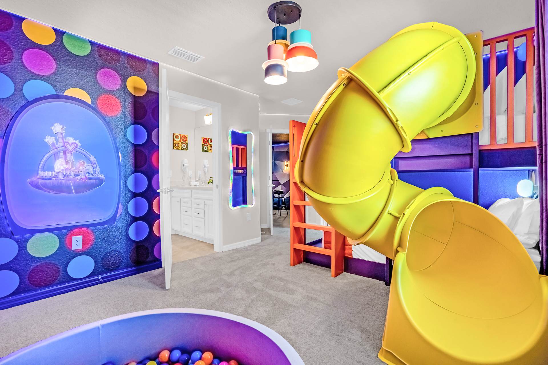 Playful kids' bedroom with a bright yellow slide attached to a purple and orange bunk bed, a wall decorated with colorful polka dots, and a ball pit. A doorway leads to an en-suite bathroom, maintaining the room's vibrant theme.