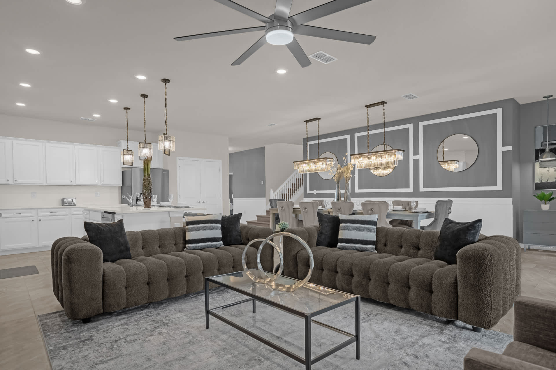 Open-concept living room with two large textured gray sofas, a glass coffee table, and a ceiling fan, flowing into a dining area with elegant chandeliers and a white kitchen in the background.
