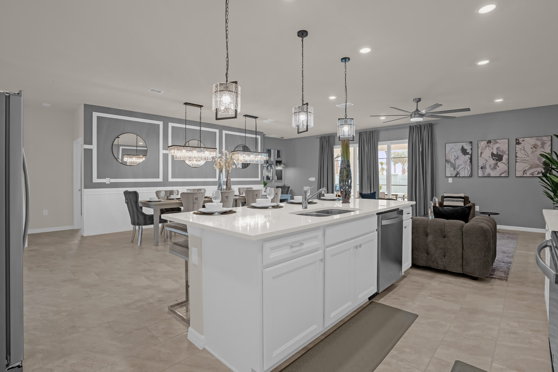An open-concept kitchen, dining, and living area featuring a white island with a double sink and modern pendant lights. The space includes a large dining table with upholstered chairs and a grey sectional sofa, all set on light tiled flooring.