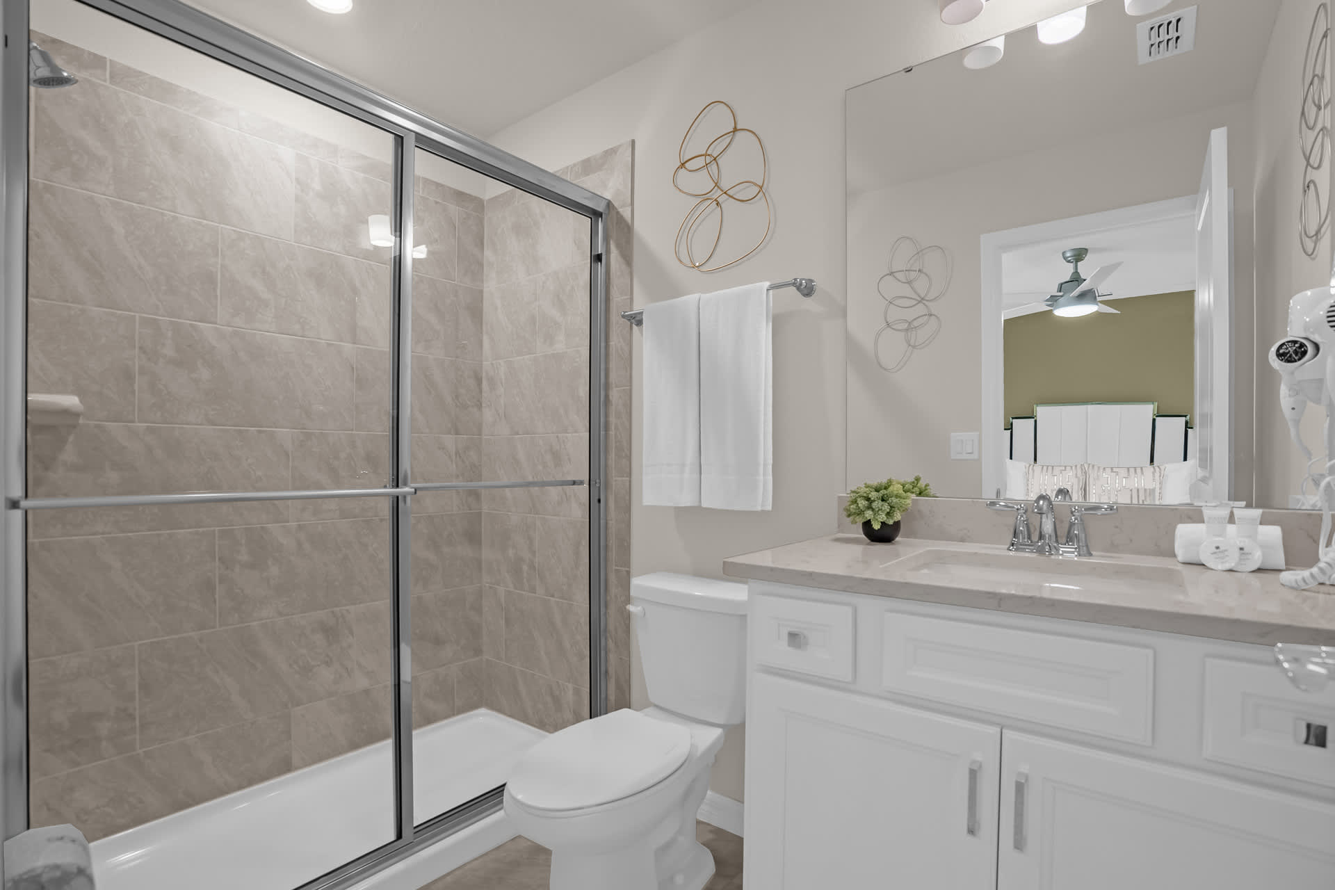 A modern bathroom with a glass-enclosed walk-in shower featuring light-colored tiles, a white vanity with a single sink, and a toilet, reflecting a ceiling fan in the mirror.