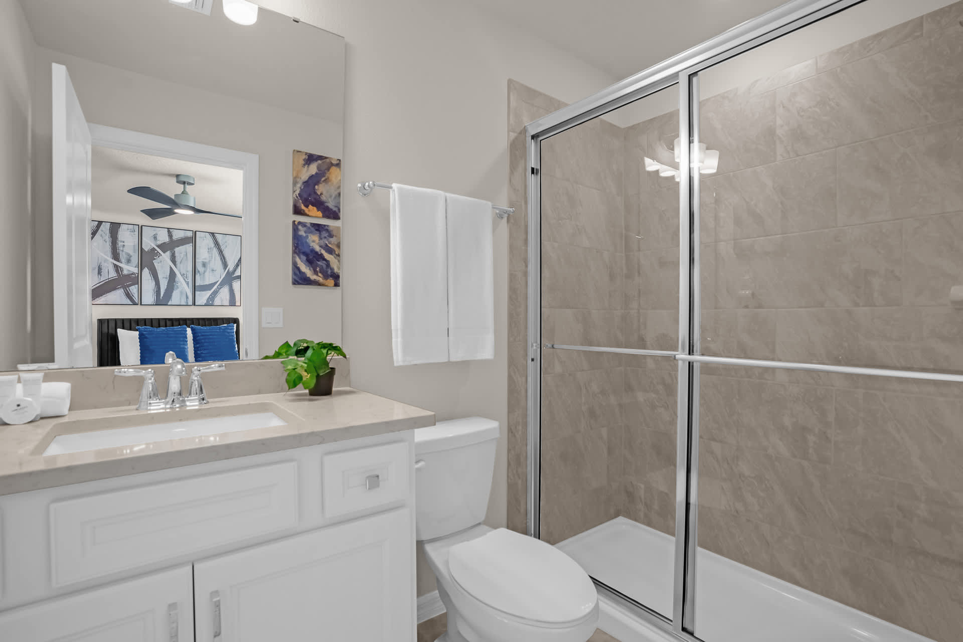 A clean bathroom featuring a glass-enclosed walk-in shower with light-colored tiles, a white vanity with a single sink, and a toilet, with decorative wall art above the toilet.