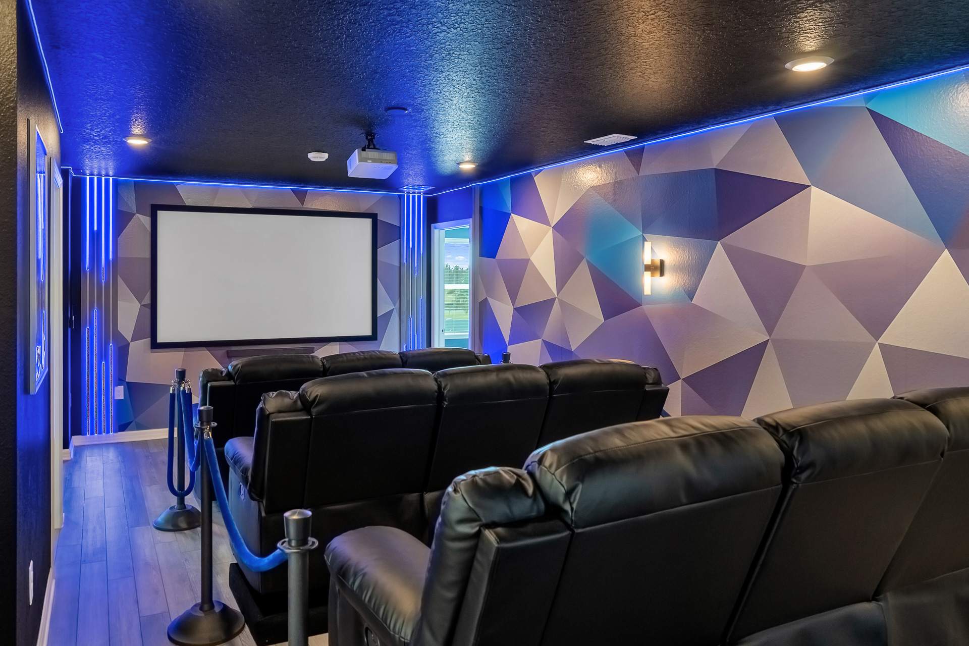 Modern home theater with rows of black leather recliners facing a large projection screen, accented by geometric blue and purple wall art and vibrant blue LED strip lighting.