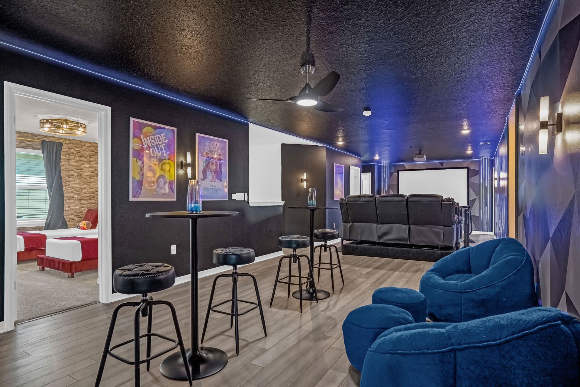 A home cinema room with a large projector screen, tiered black leather recliners, and a small bar area with high-top tables and stools, illuminated by blue LED strip lighting.