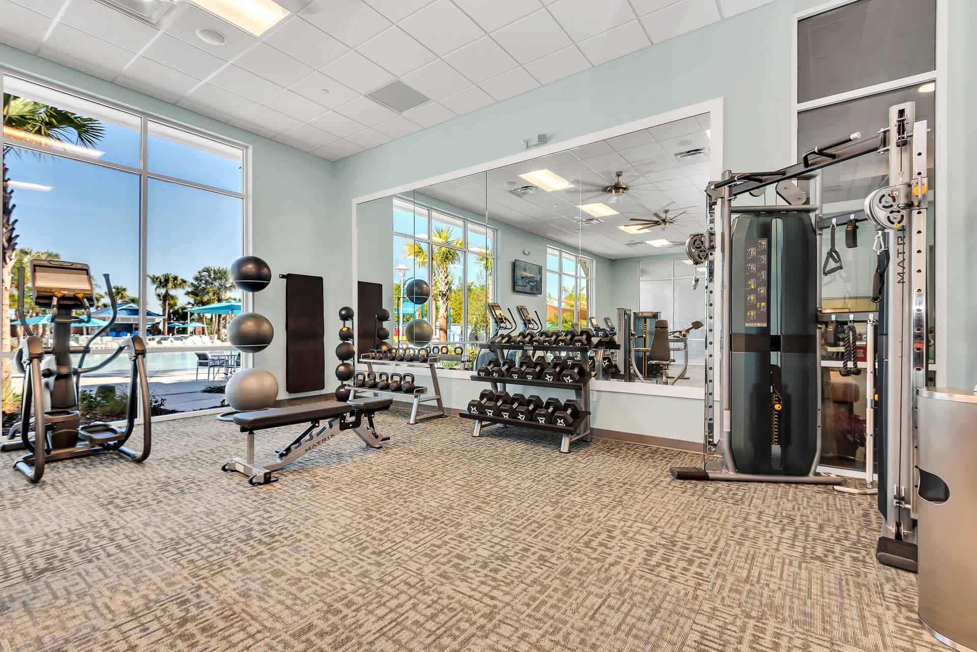 A well-equipped fitness center includes a variety of free weights, benches, and strength training machines, with a large mirror reflecting the room and outdoor views through expansive windows.