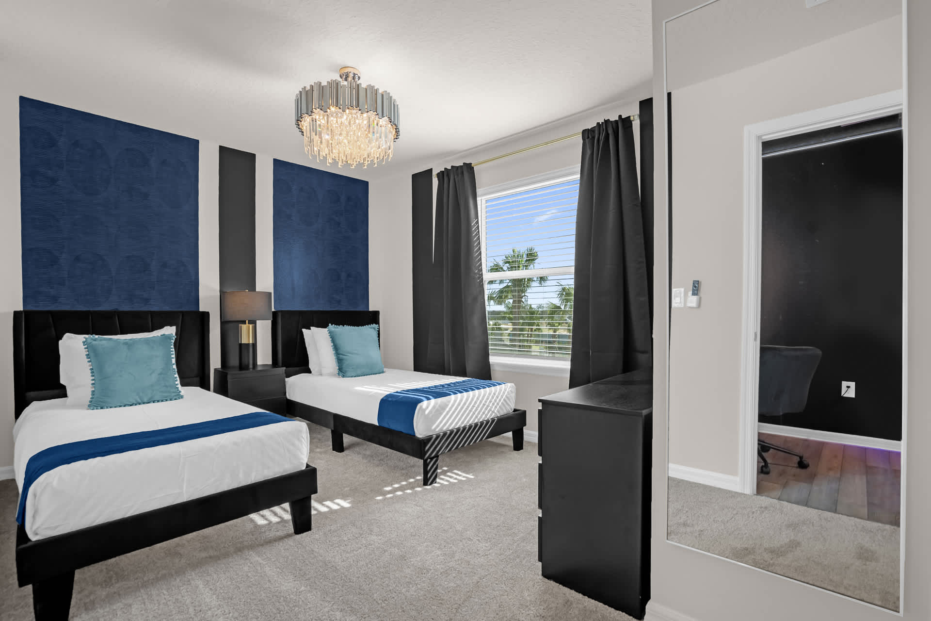 A bright bedroom designed with two twin beds, each with a black frame and blue accents, a modern chandelier, and a window with dark curtains, featuring a blue and black accent wall.