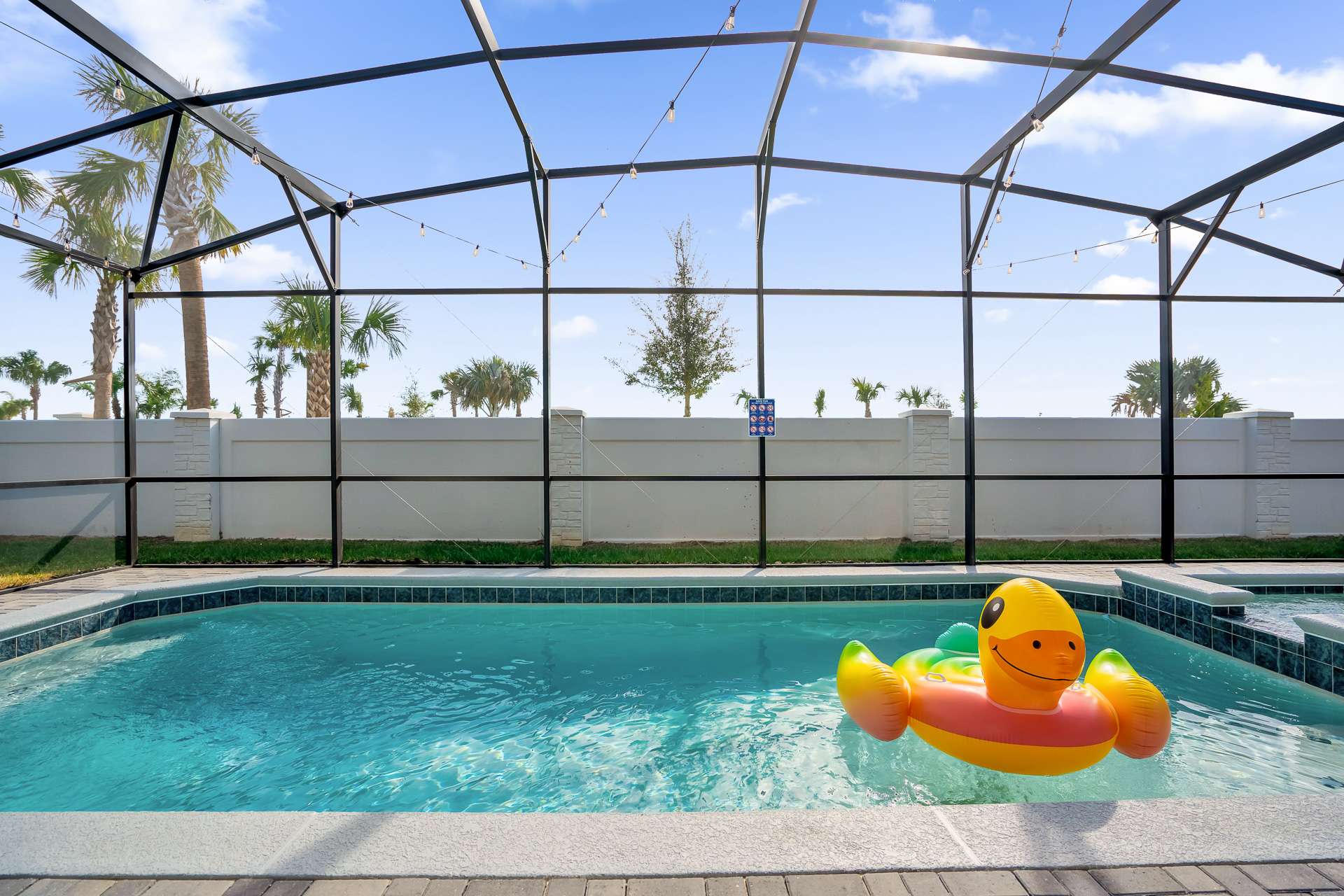Screened-in private swimming pool with clear blue water and an inflatable duck float, surrounded by a paved deck and a low wall, with palm trees visible beyond the enclosure.