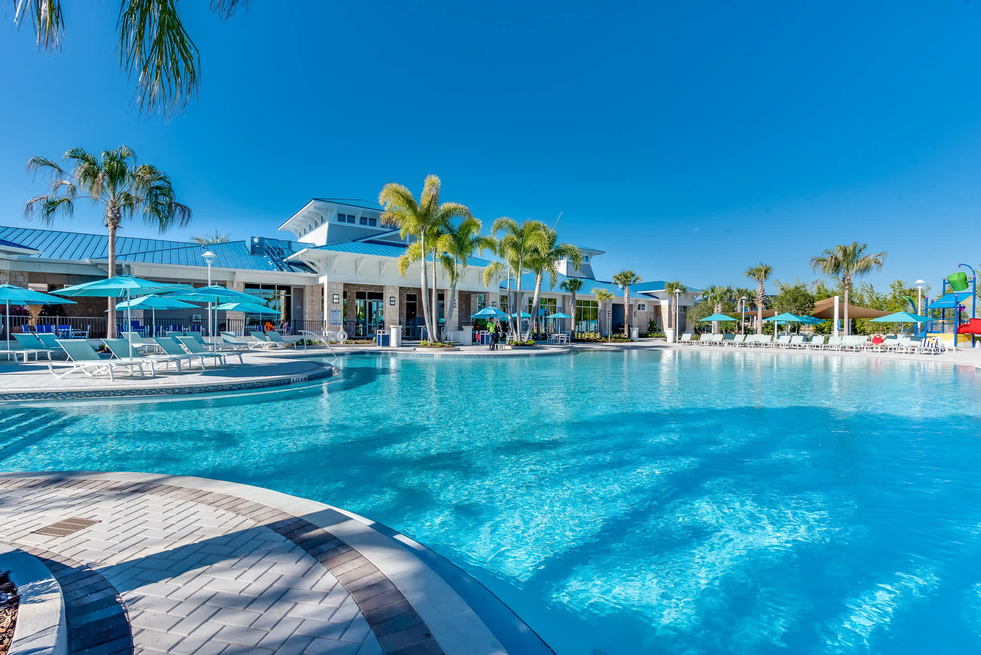 Expansive outdoor swimming pool with clear blue water, surrounded by light-colored pavers, numerous lounge chairs with blue umbrellas, and tall palm trees, with a modern resort building in the background under a bright blue sky.
