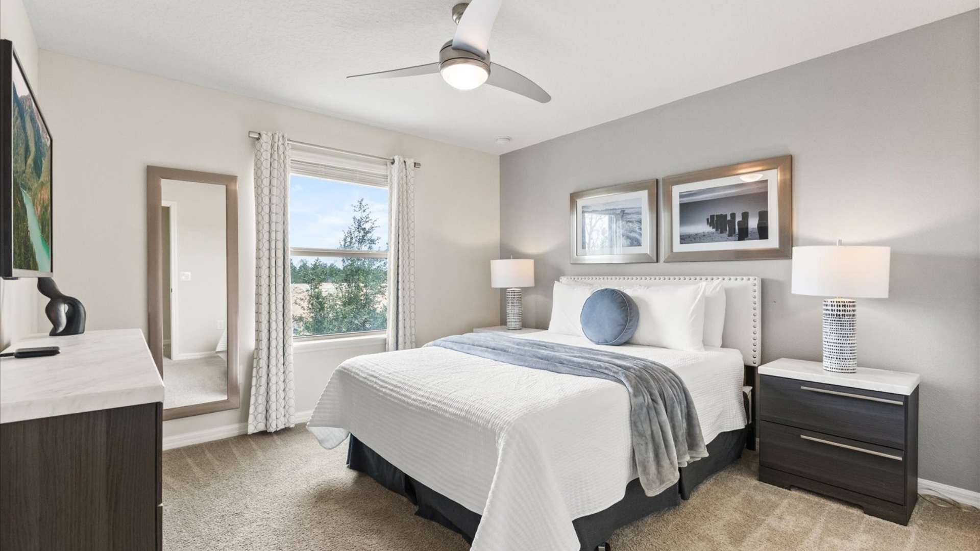 Cozy bedroom with a queen-sized bed featuring a white quilted comforter and a blue throw, flanked by dark nightstands with modern lamps, and a large window with patterned curtains.