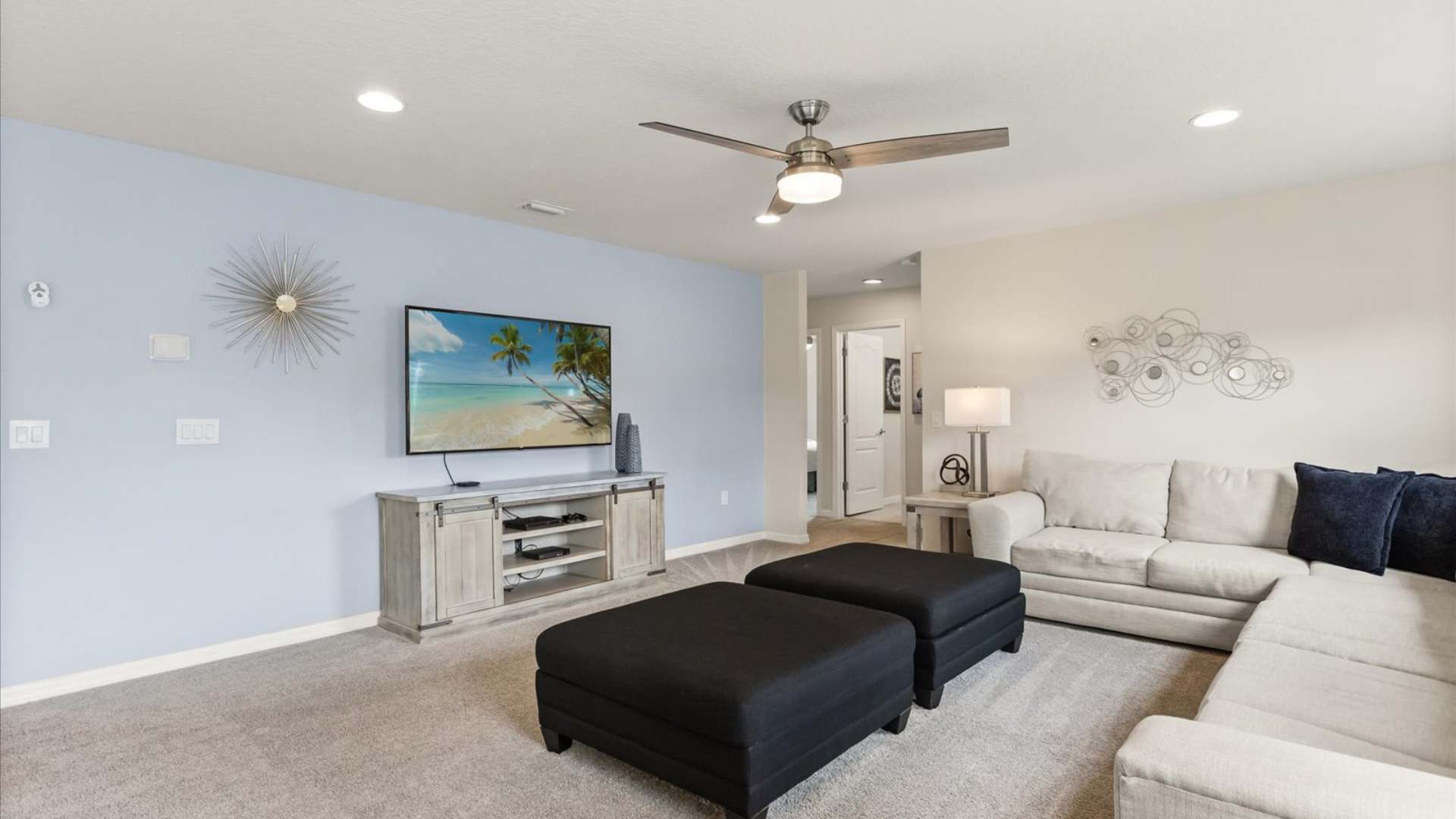 Upstairs media room with light gray carpet, a large sectional sofa, two black ottomans, and a wall-mounted TV above a light wood console, featuring a light blue accent wall.