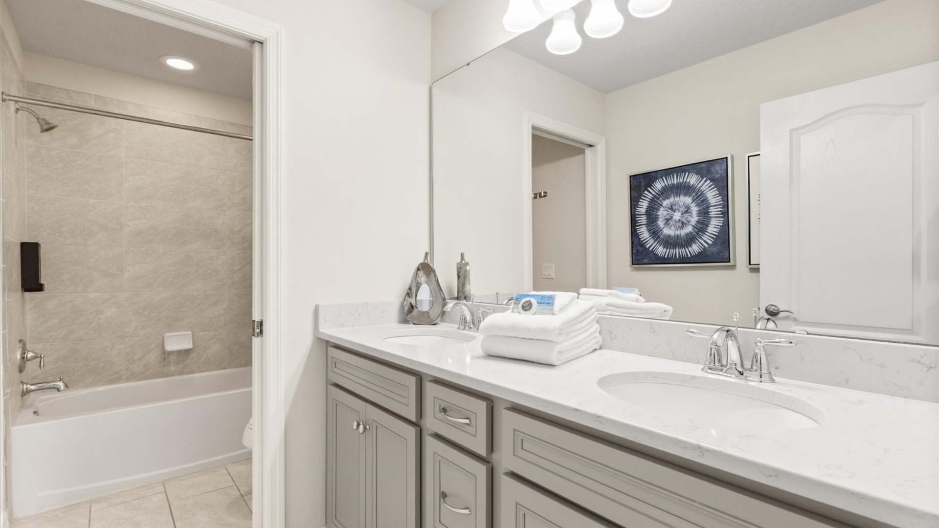 Modern bathroom featuring a double vanity with light gray shaker-style cabinets and white quartz countertops, a large mirror, and a shower-tub combo with light gray tiled walls.