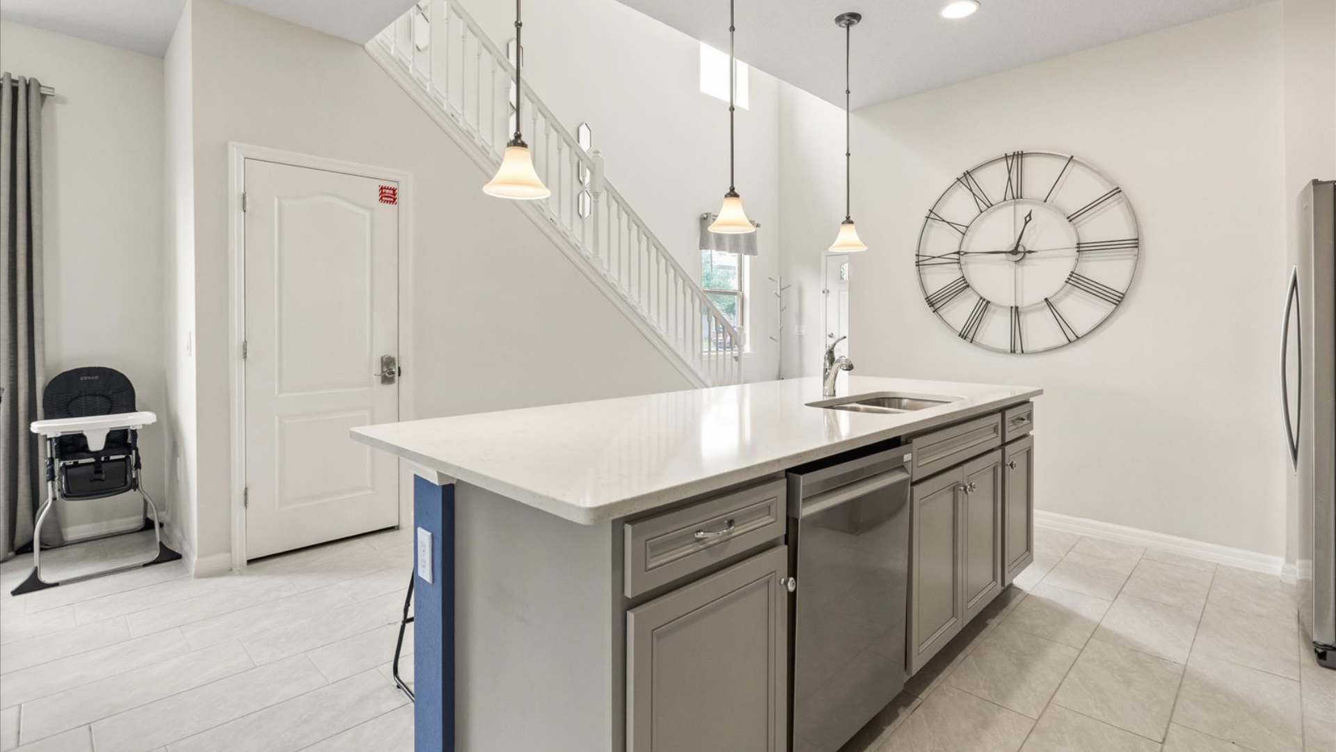 Kitchen island with a double sink and white quartz countertop, featuring pendant lights overhead, and a view of the grand staircase with white railings leading upstairs.