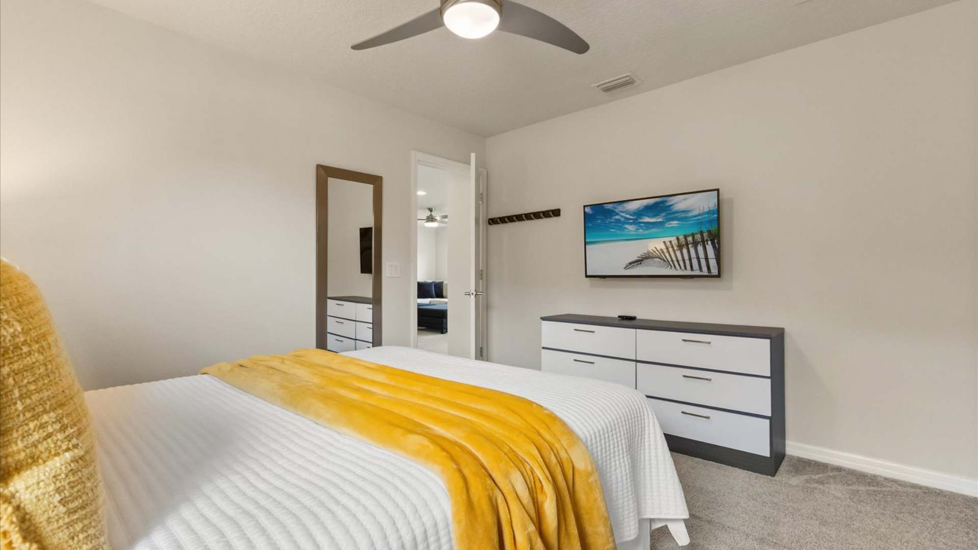 View of a bedroom showing a white and gray dresser with a wall-mounted TV, a full-length mirror, and a bed with yellow accents, all on a light-colored carpet.