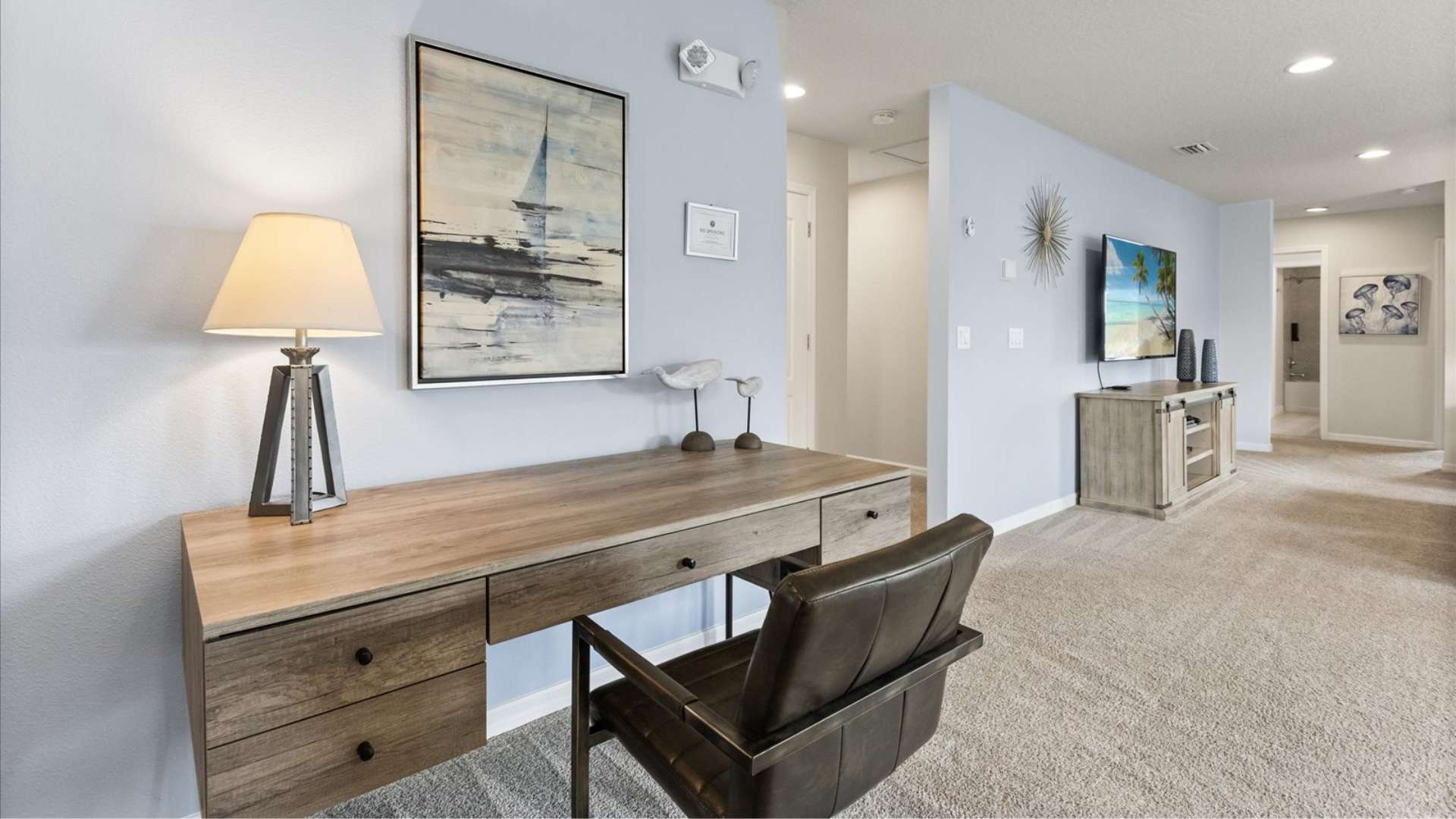 Dedicated office space with a rustic wooden desk, a dark leather chair, and a modern table lamp, set against a light blue wall with abstract artwork, with a TV and console visible in the background.