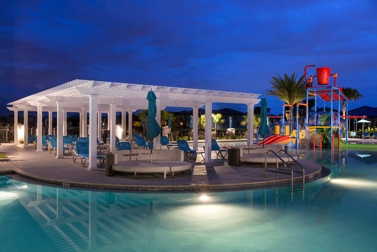 Illuminated resort pool area at dusk, featuring a large swimming pool with underwater lights, a white pergola with seating, and a colorful water play structure in the background, all under a dark blue sky.