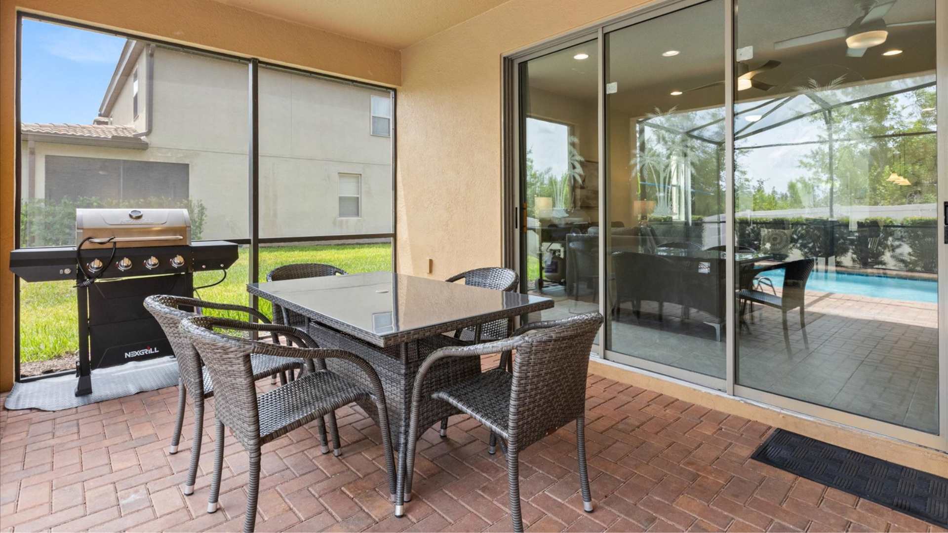 Covered outdoor patio with a BBQ grill and a glass-top dining table with wicker chairs, accessible from the living room via large sliding glass doors. The screened pool area is visible beyond.