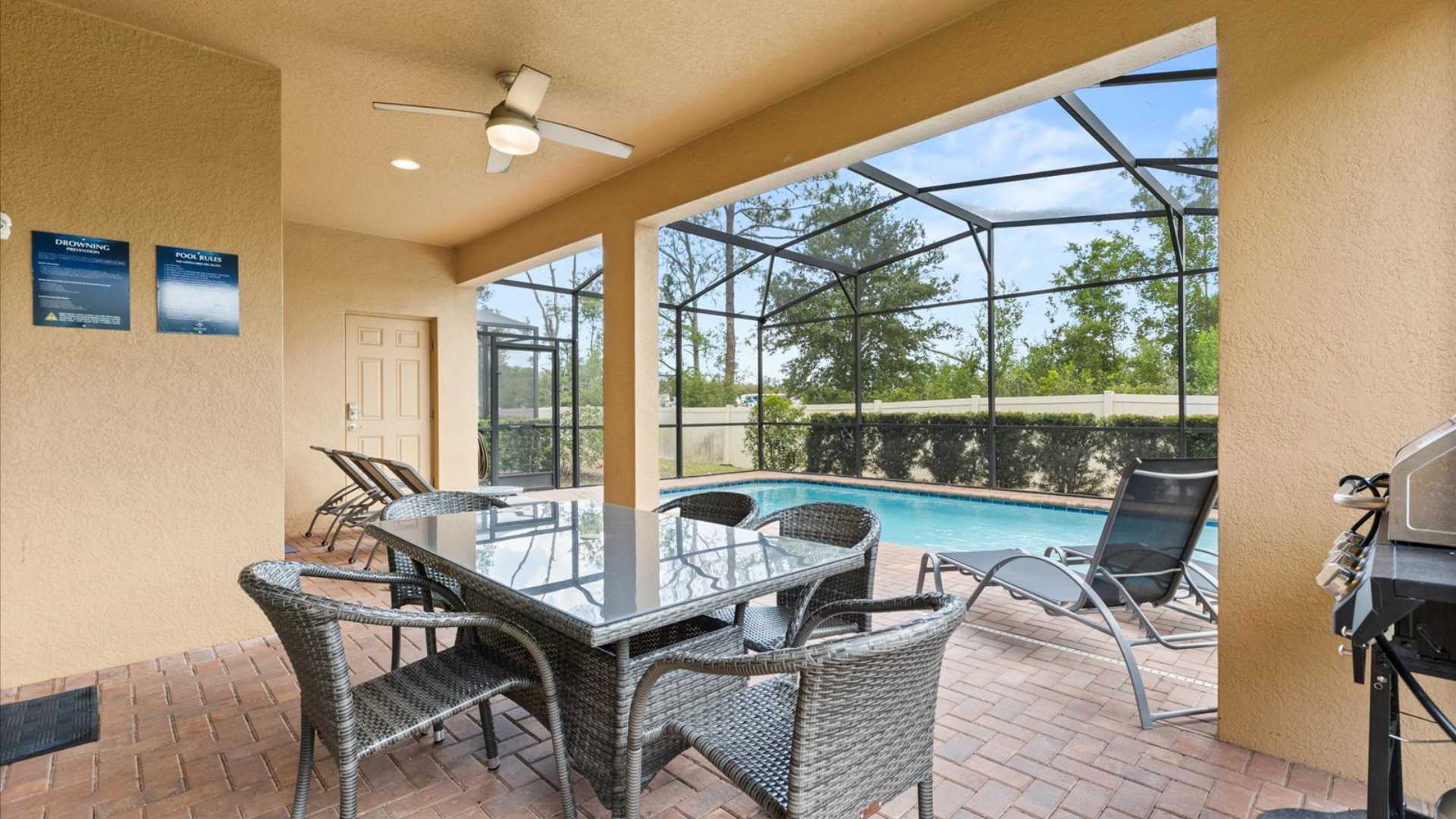 Covered outdoor patio area with a glass-top dining table and wicker chairs, adjacent to a screened-in private pool. A ceiling fan provides comfort, and lounge chairs are visible by the pool.