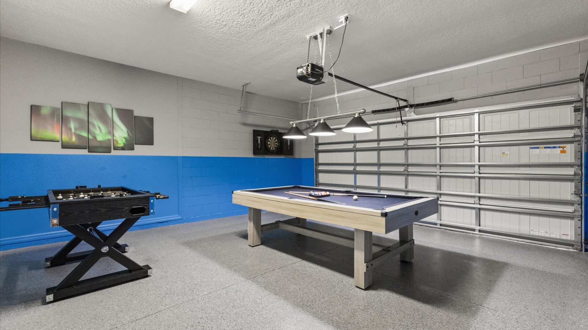 Wide shot of the game room, showcasing a foosball table, a pool table with overhead lighting, and a dartboard. The garage door is visible, confirming its conversion into a recreational space.