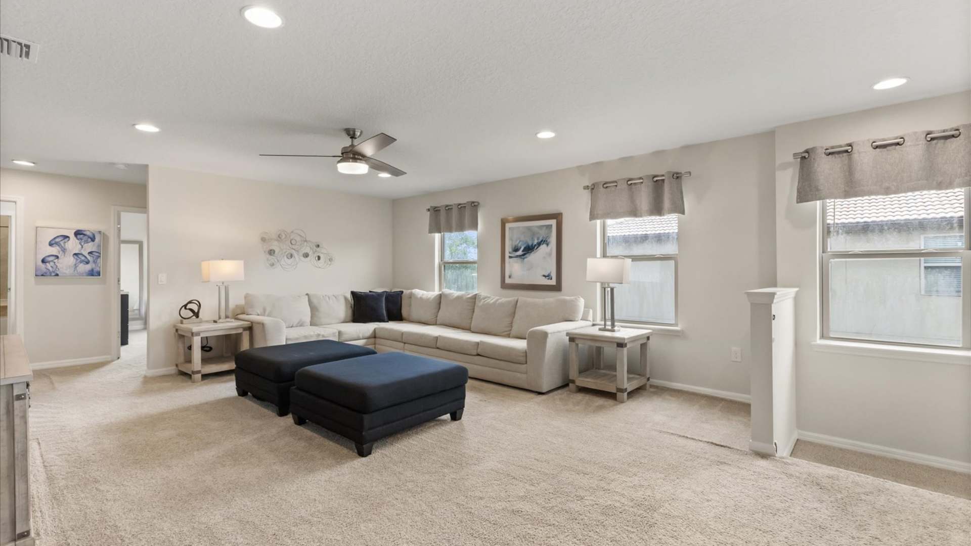 Bright living room featuring a large cream sectional sofa with navy throw pillows, two navy ottomans, and light beige carpet, illuminated by recessed lighting and natural light from multiple windows with grey valances.