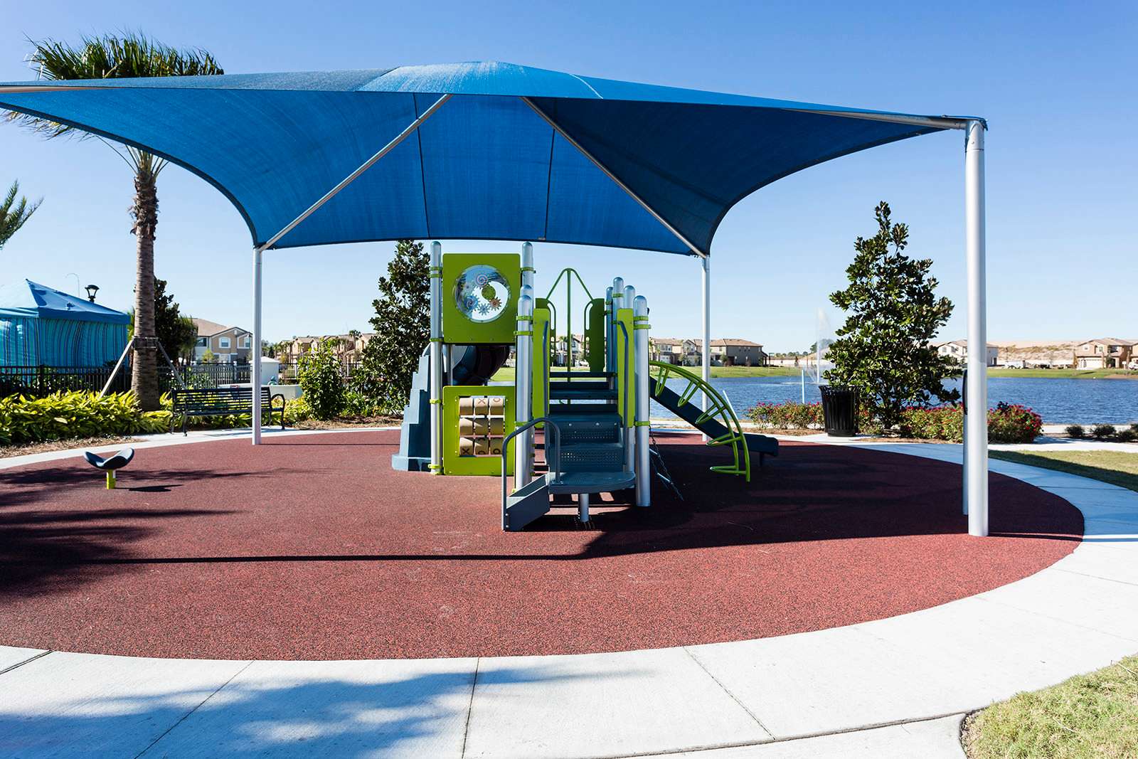 Vibrant children's playground with a large blue shade canopy, featuring green and gray play structures on a red rubberized surface, surrounded by green landscaping and palm trees.