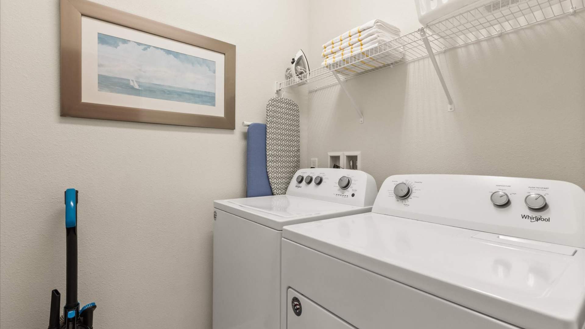 Functional laundry room equipped with a white top-loading washing machine and a front-loading dryer, a wall-mounted shelf with towels, an ironing board, and a framed artwork on the wall.