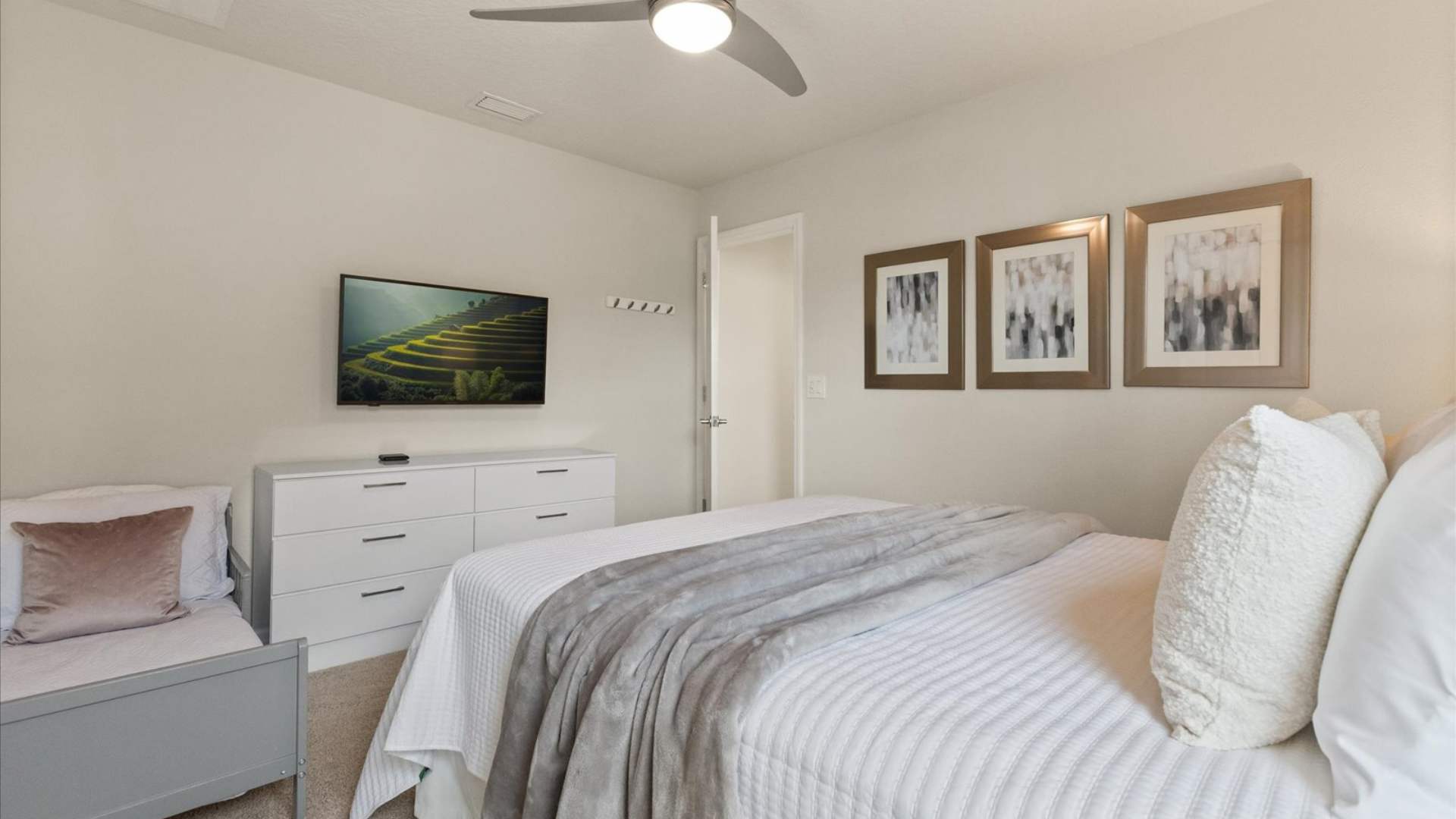 Cozy bedroom with a white quilted bed, a grey throw blanket, and a small grey daybed, featuring a white dresser with a wall-mounted TV above it, and three framed abstract artworks on the wall.
