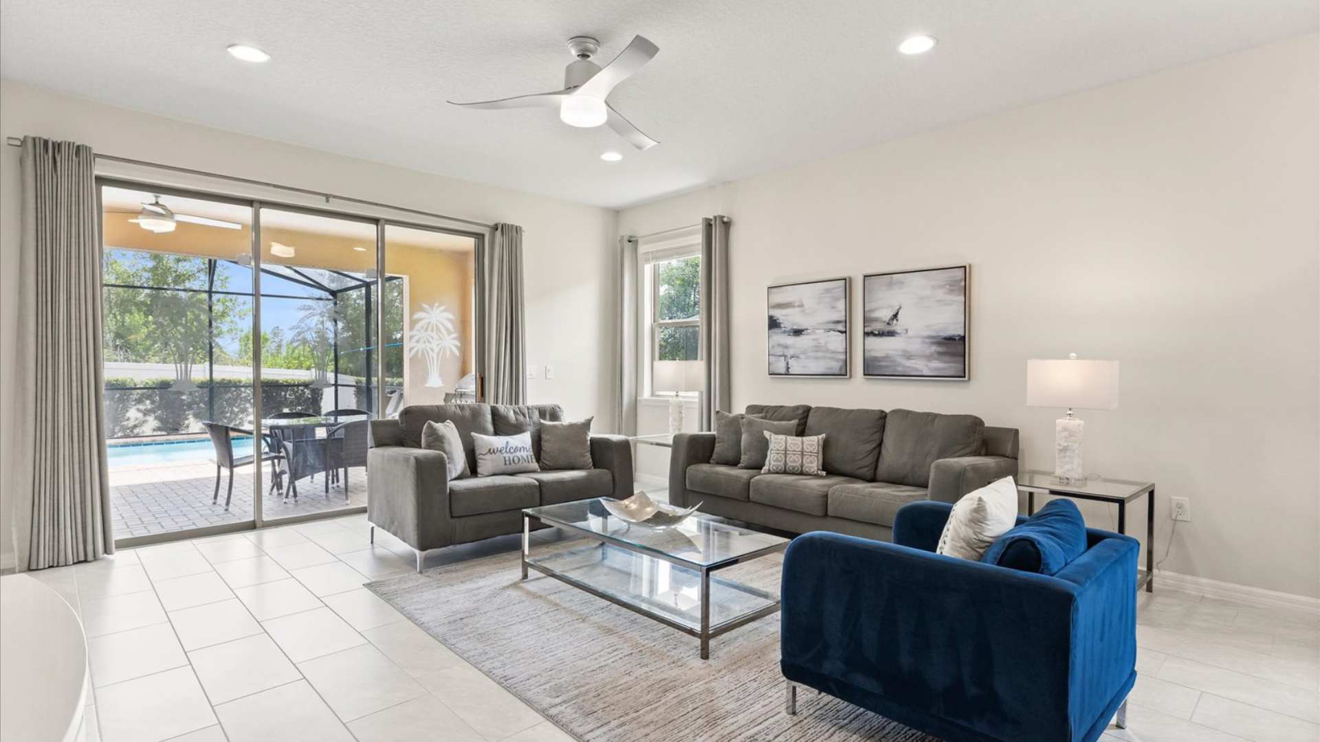 Bright living room with light tile flooring, two gray sofas, a blue accent chair, and a glass coffee table, featuring large sliding doors opening to a screened patio with a pool.