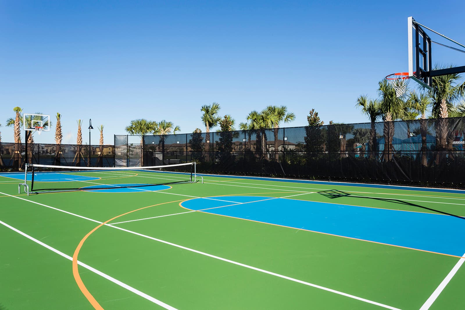 Multi-sport court with a vibrant green and blue surface, featuring a basketball hoop and a volleyball net, enclosed by a black fence and lined with palm trees under a clear blue sky.