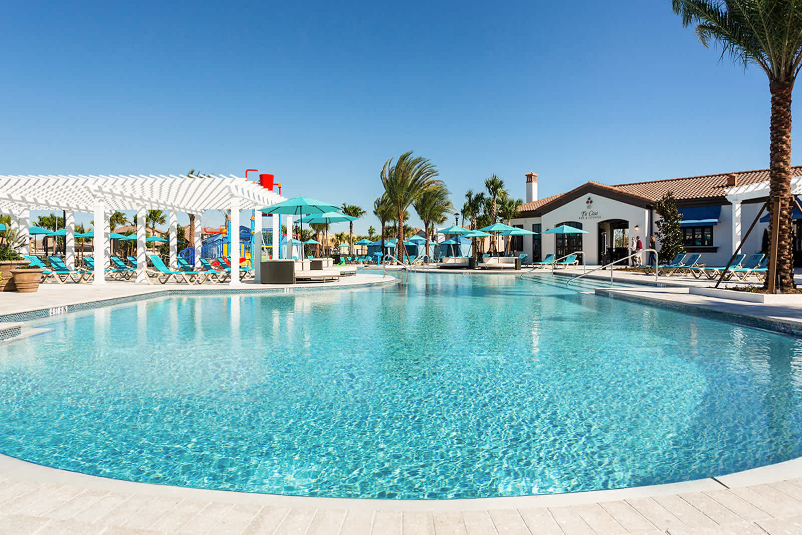 Expansive resort-style swimming pool with clear blue water, surrounded by lounge chairs, teal umbrellas, and a white pergola, with a clubhouse building in the background under a bright blue sky.