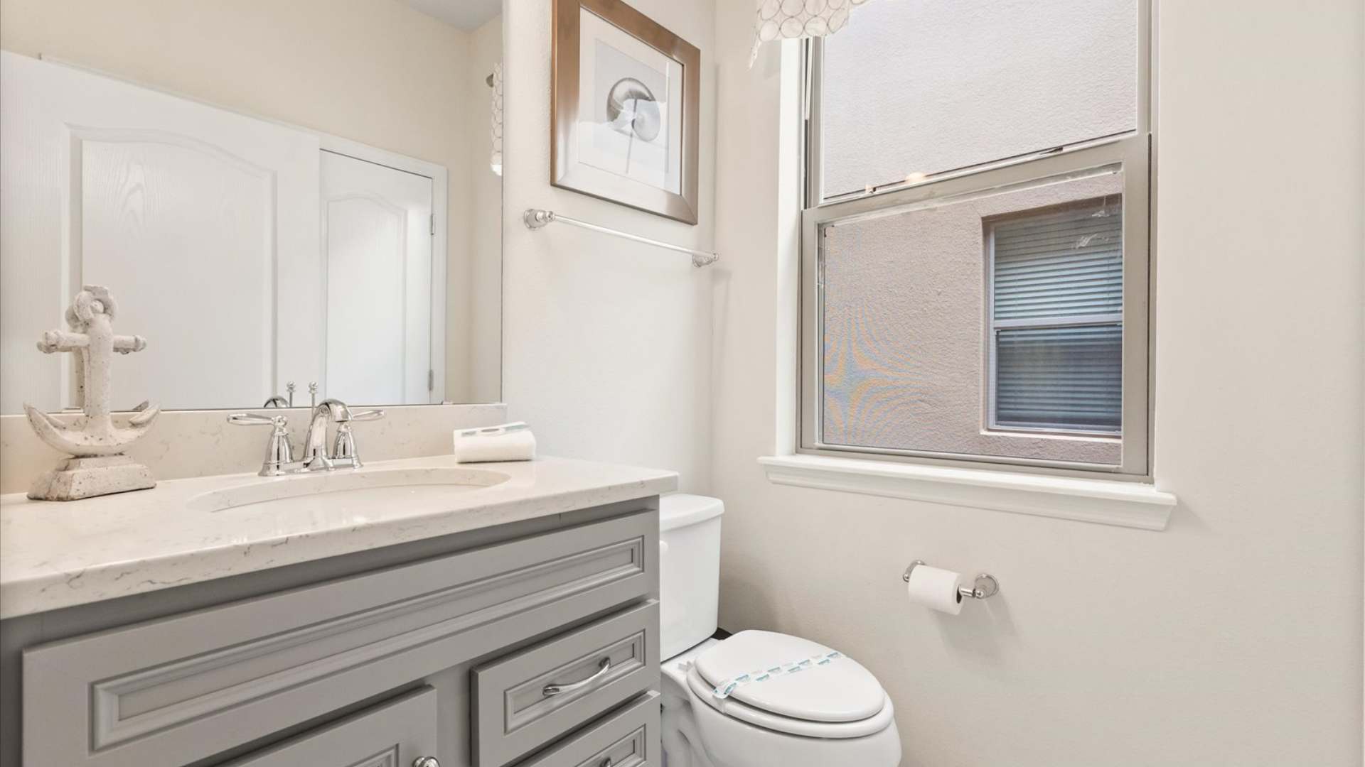 Compact bathroom with a single gray vanity, white countertop, and a toilet, featuring a window with blinds and a decorative anchor statue on the counter.