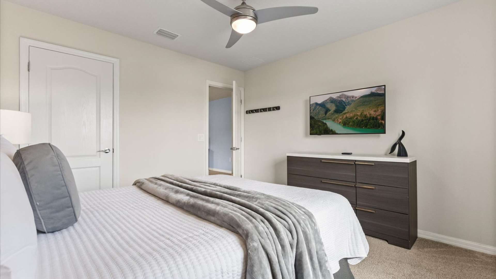Bright bedroom featuring a queen-sized bed with a white comforter and gray throw, a dark wood dresser with a wall-mounted TV, and a light-colored carpeted floor.