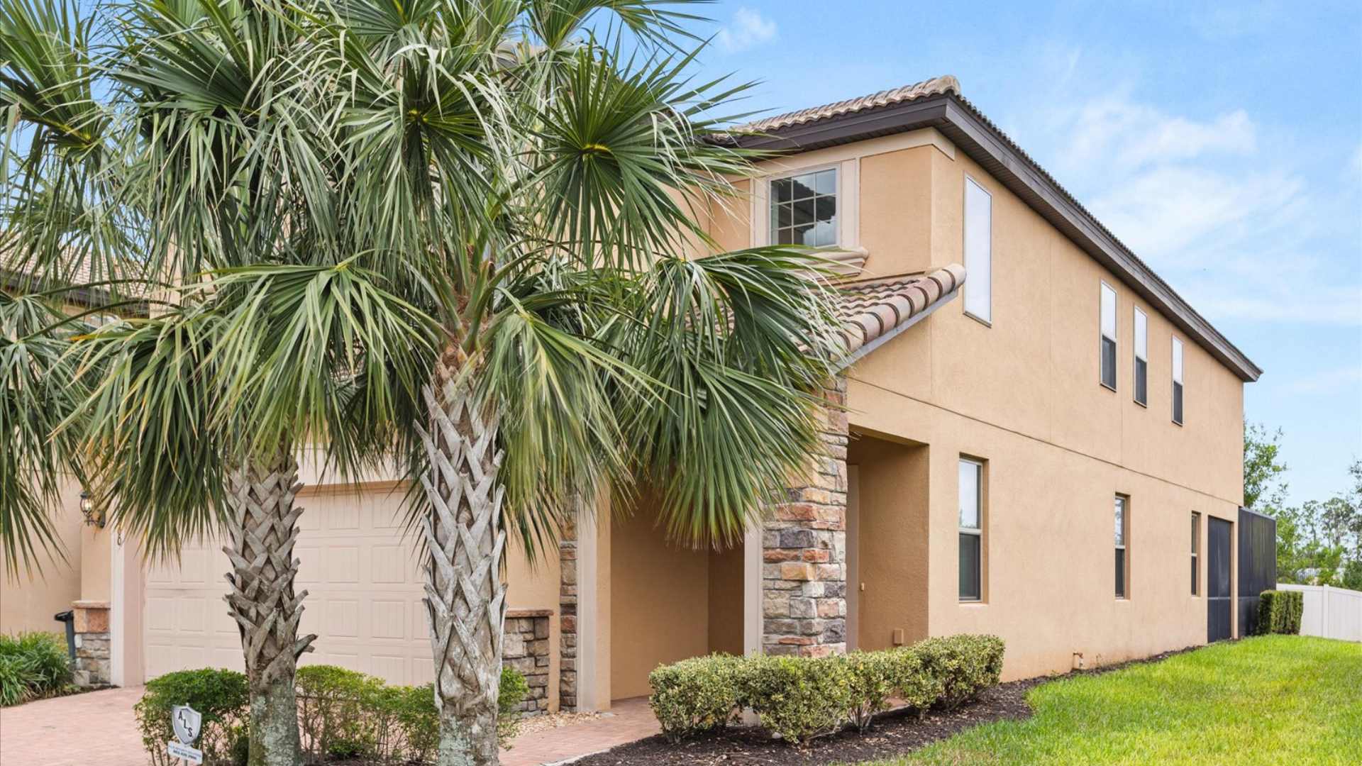 The side view of a two-story house showcasing warm stucco walls, decorative stone column accents, and a tiled roof, with a paved walkway leading to the front entrance, flanked by green shrubs and palm trees.