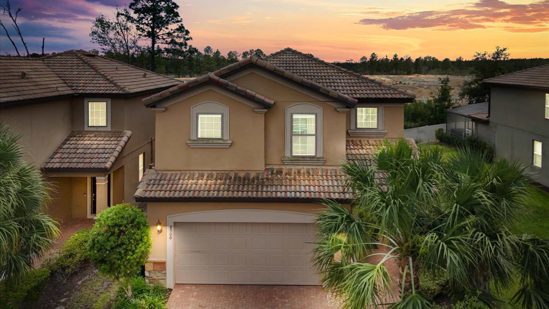 The front exterior of a two-story house with a tiled roof and an attached two-car garage, bathed in the warm hues of a sunset sky, surrounded by lush palm trees and landscaping.