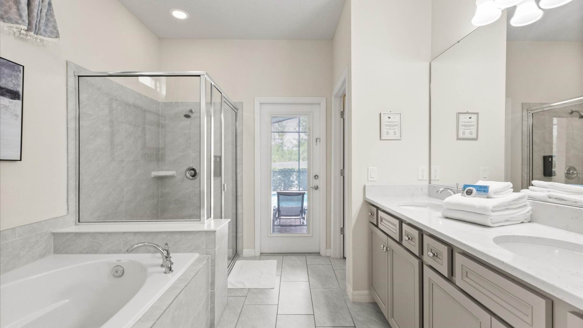 Spacious bathroom with a large soaking tub and a glass-enclosed walk-in shower with grey tile, featuring light grey floor tiles and a door leading to an outdoor area, suggesting pool access.