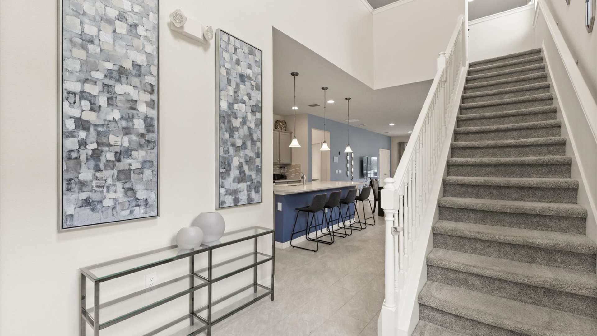 Bright foyer area with light tile flooring, featuring a glass console table with decorative vases, modern abstract artwork, and a carpeted staircase leading to the upper floor.